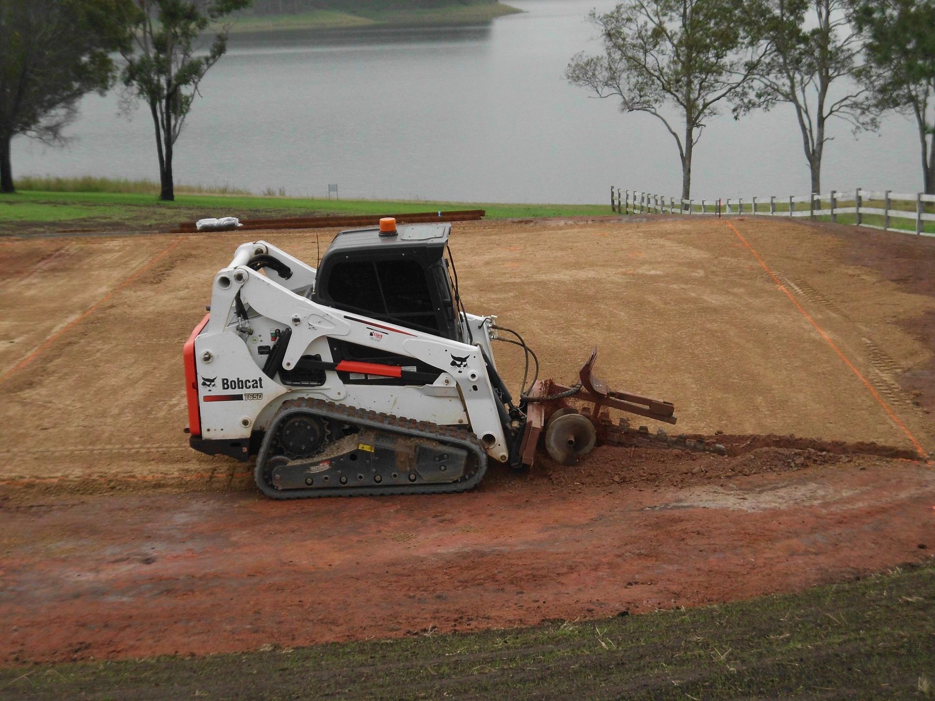 White Bobcat Skid-Steer with A Tiller Attachment Leveling Red Dirt by A Body of Water — Evans Earthmoving Contractors In Malanda, QLD 