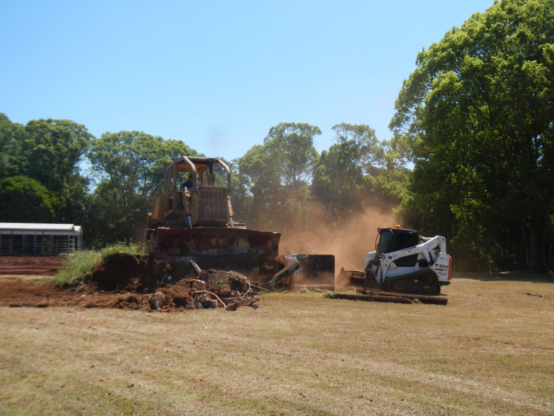 A Small Bulldozer and Skid Steer Demolishing a Concrete Structure in A Grassy Field — Evans Earthmoving Contractors In Malanda, QLD 