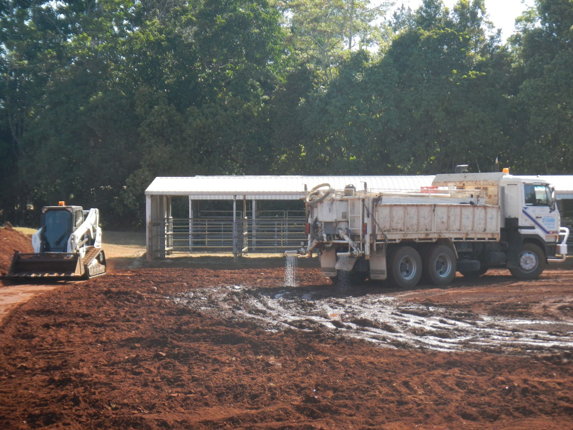 Construction Site: A Truck Depositing Mud as A Skid Steer Loader Prepares the Ground, Near a White Building — Evans Earthmoving Contractors In Malanda, QLD 