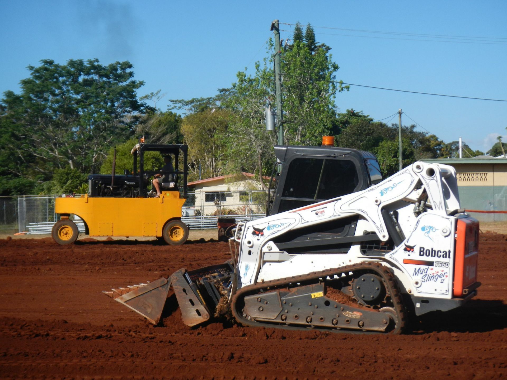 White skid-steer loader pushing dirt on a construction site beside a yellow forklift on red soil — Evans Earthmoving Contractors In Malanda, QLD 