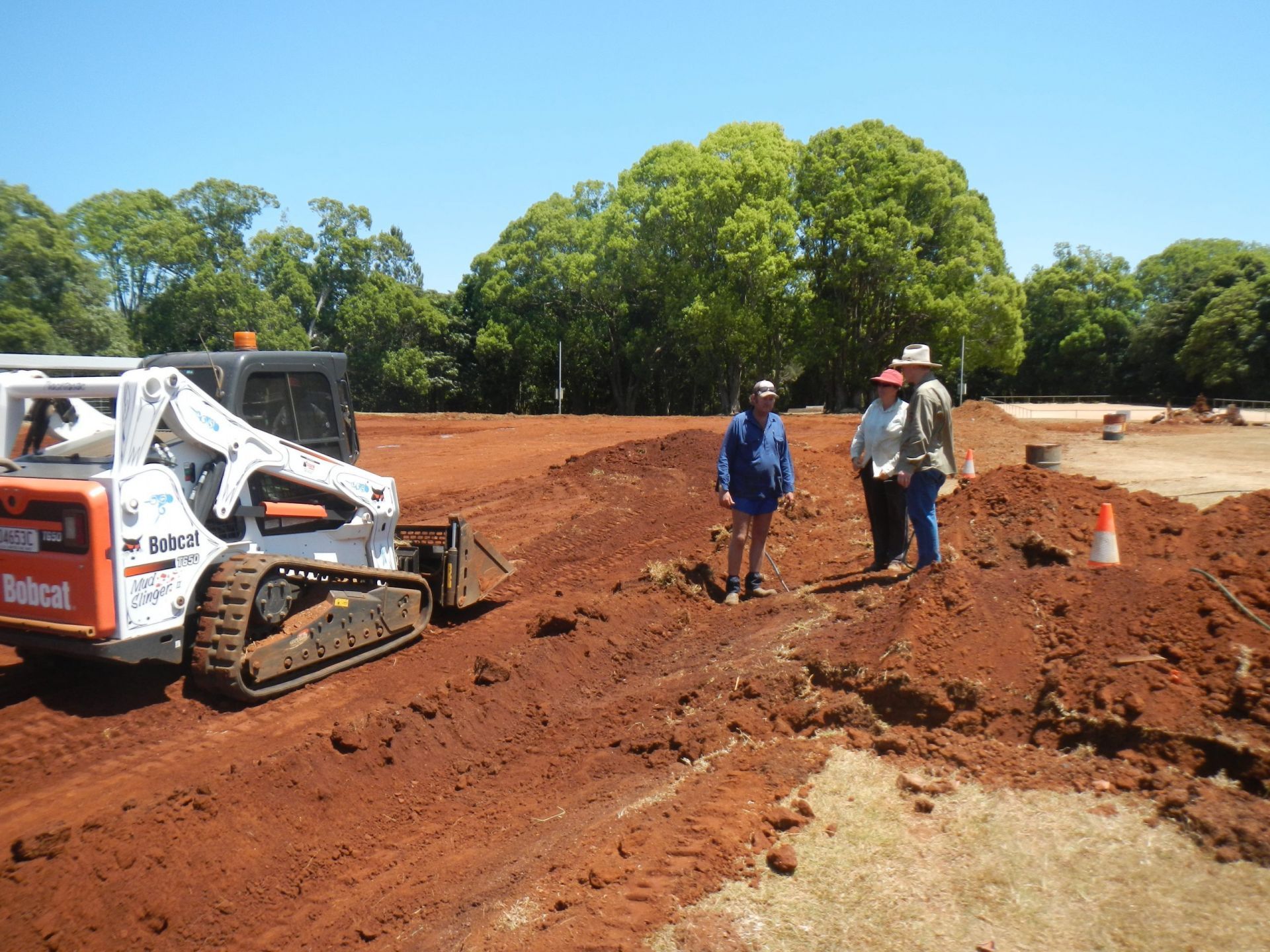 Bobcat Excavator and Three People on A Red Dirt Construction Site Under a Blue Sky, Trees in Background — Evans Earthmoving Contractors In Malanda, QLD 
