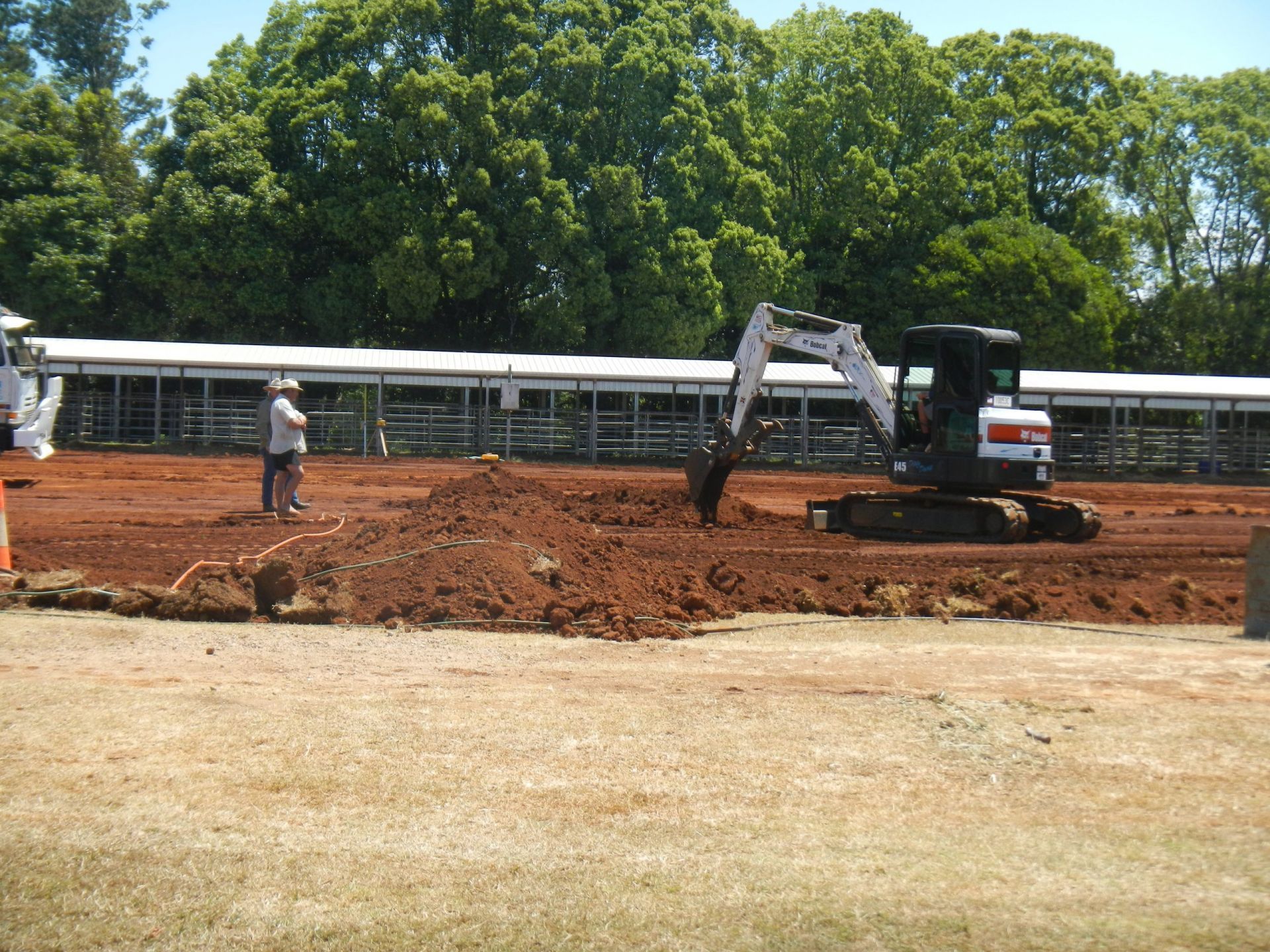 A Small Excavator Digs Into a Dirt Area with A Person Watching, Stalls in The Background, Trees Above — Evans Earthmoving Contractors In Malanda, QLD 