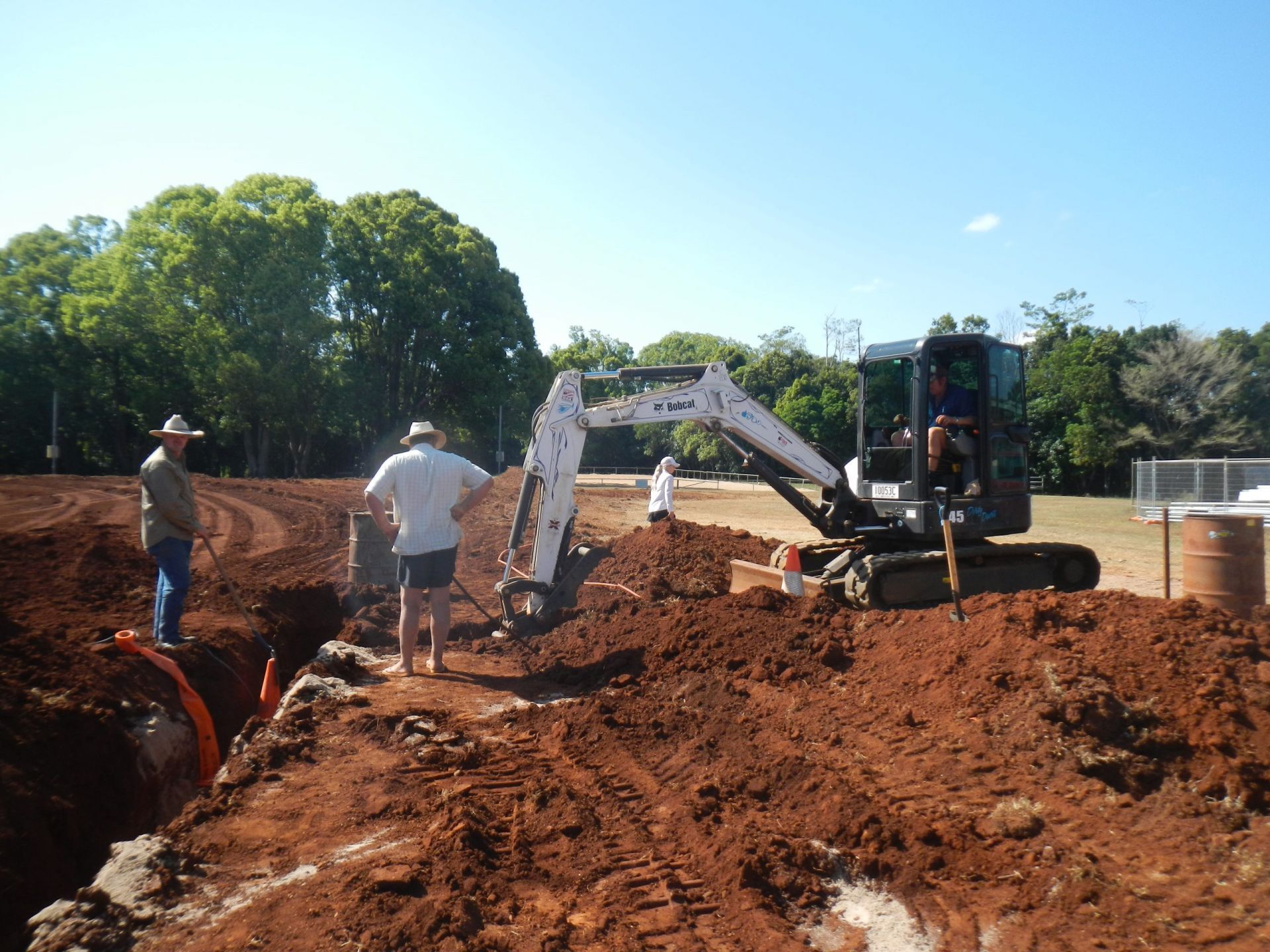 Two Men Watch an Excavator Digging in Reddish Dirt Near a Tree Line and Building — Evans Earthmoving Contractors In Malanda, QLD 