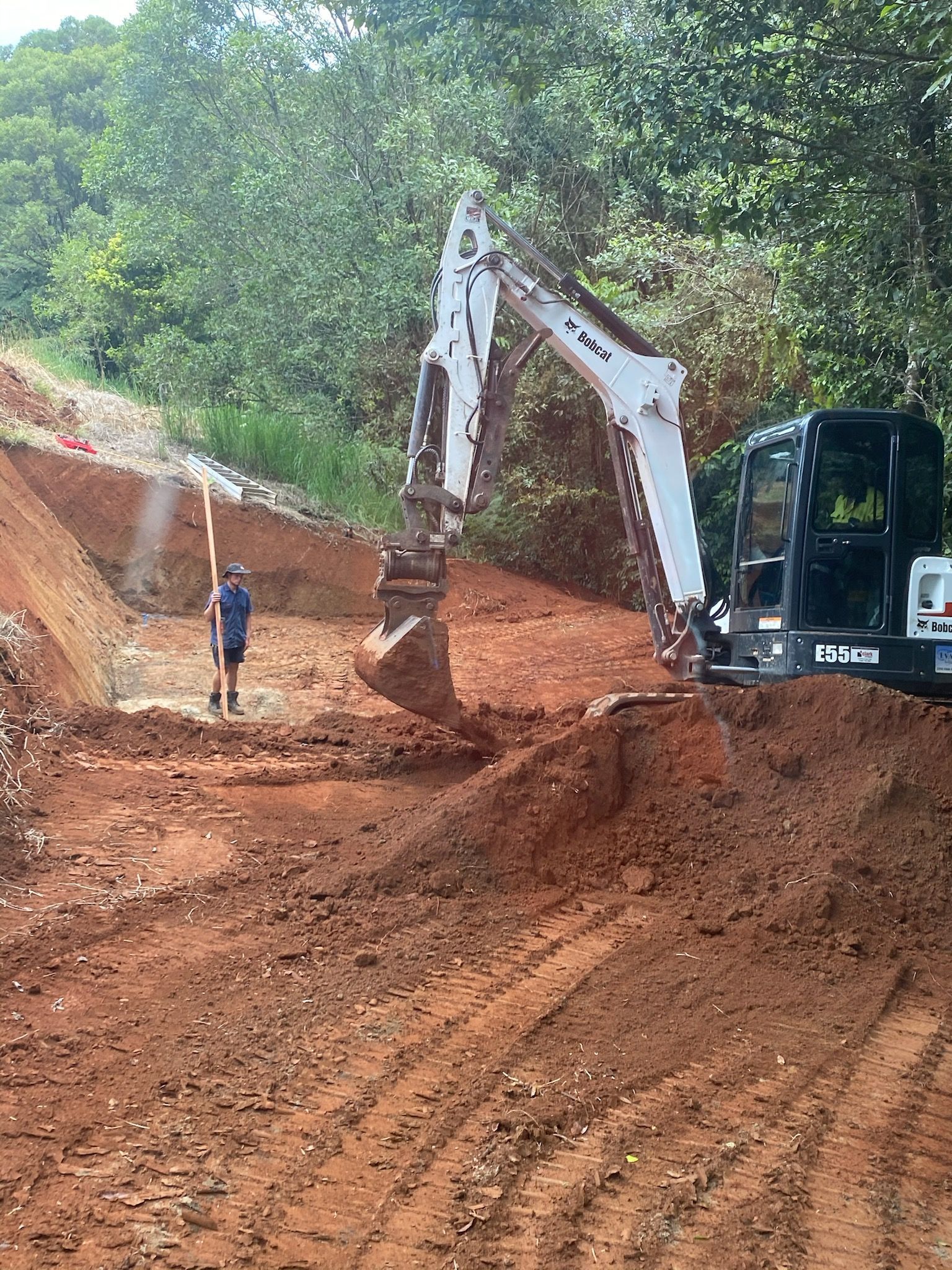 Excavator Digging in Red Soil with A Person Surveying, Forest Background — Evans Earthmoving Contractors In Malanda, QLD 