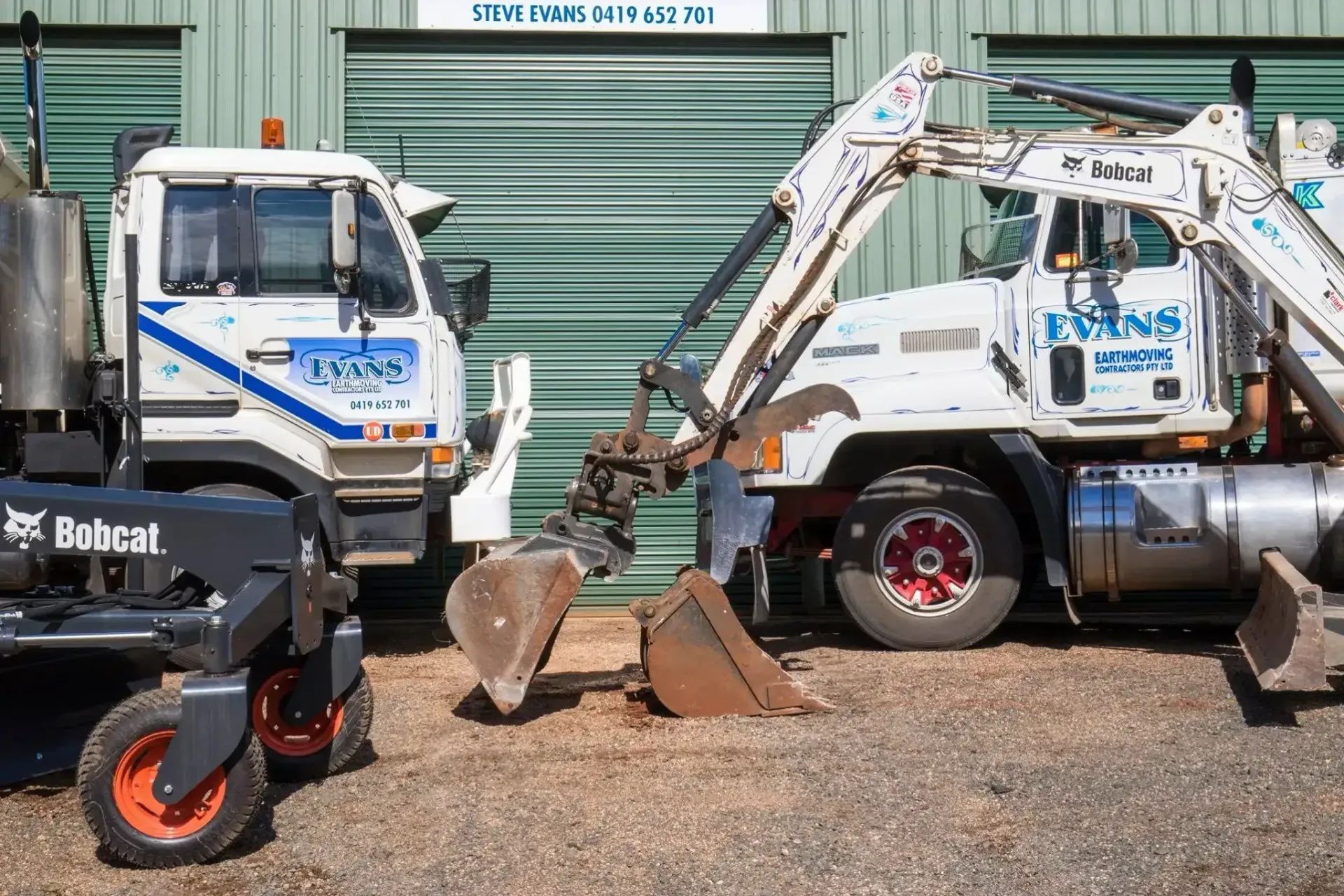 Man Stands Between Two White Trucks with Blue Logos, in Front of A Green Building — Evans Earthmoving Contractors In Malanda, QLD 