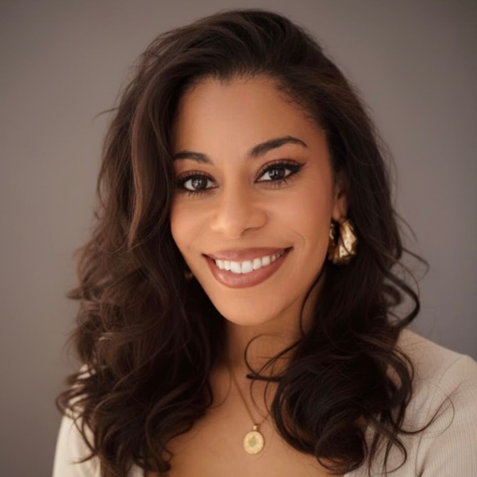 Smiling woman with wavy dark hair wearing a beige top and gold jewelry against a neutral background