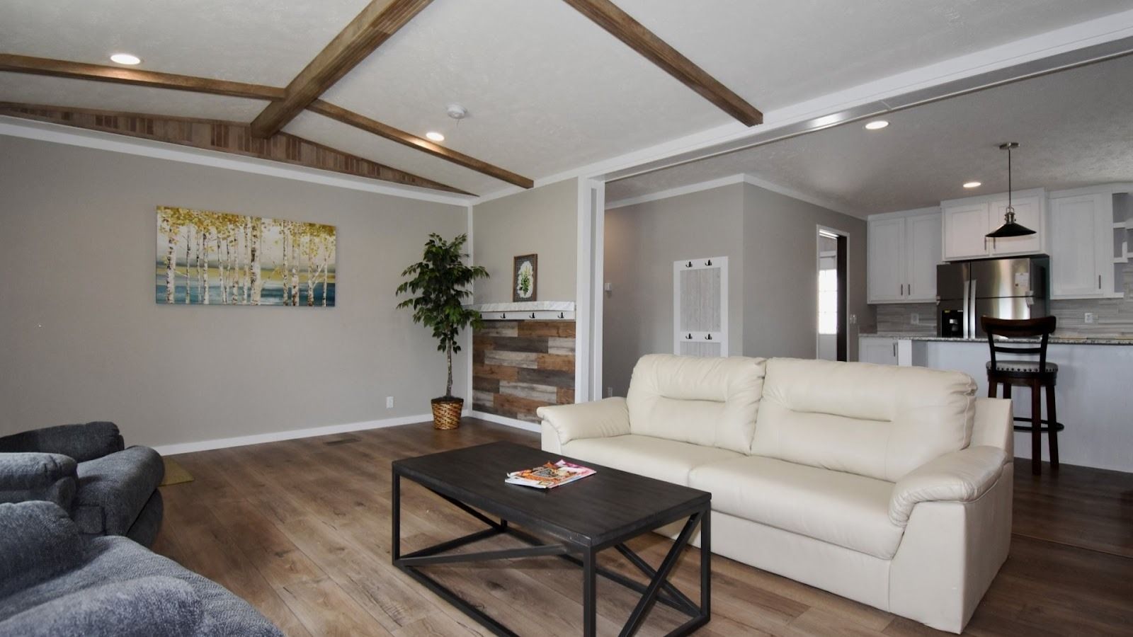 Spacious modern living room with beige leather sofa, dark coffee table with magazines, gray upholstered chairs, wooden ceiling beams, and a view into a white kitchen with stainless steel appliances