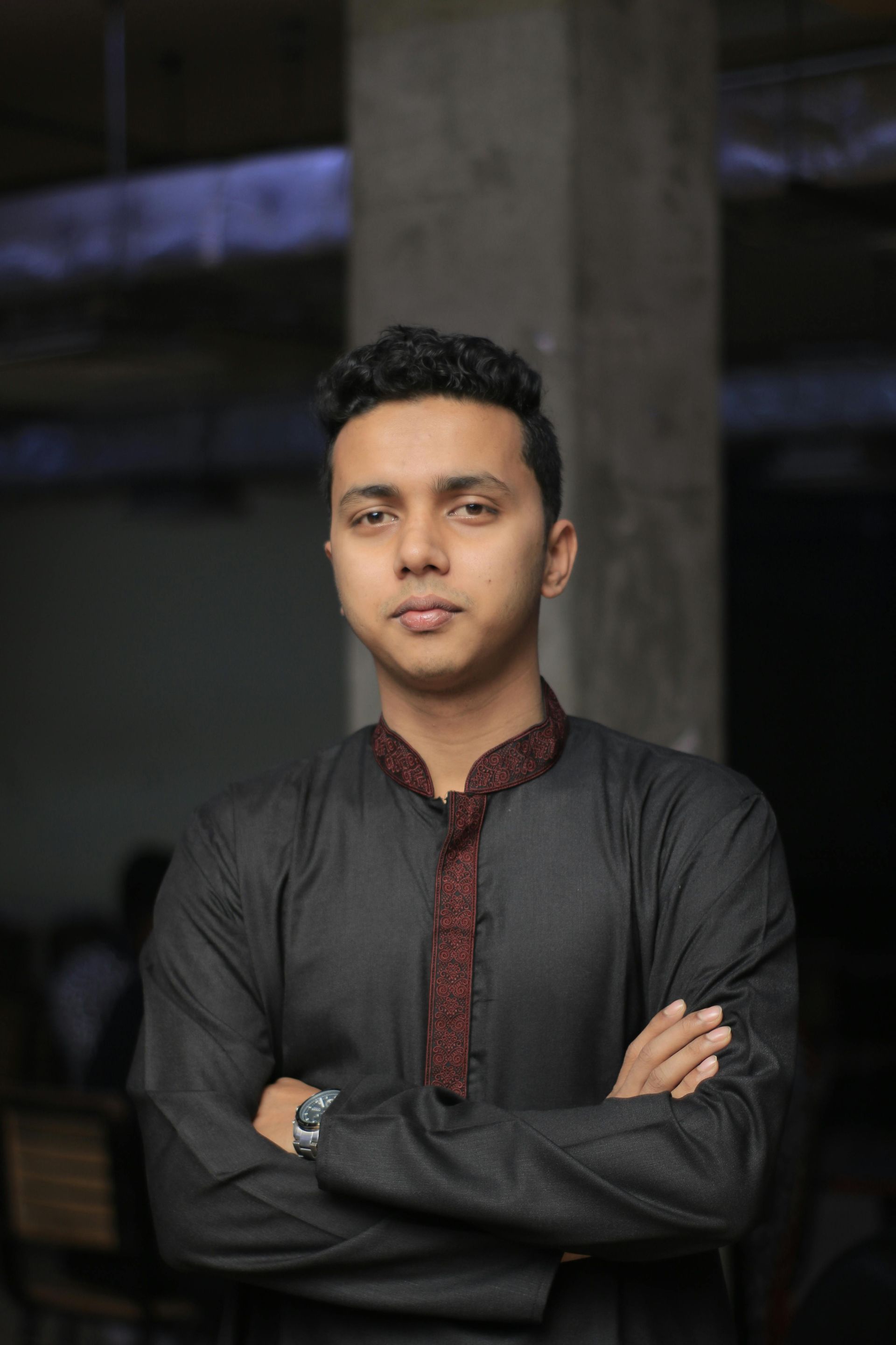 Person in a dark traditional shirt stands with arms crossed against a blurred indoor background.