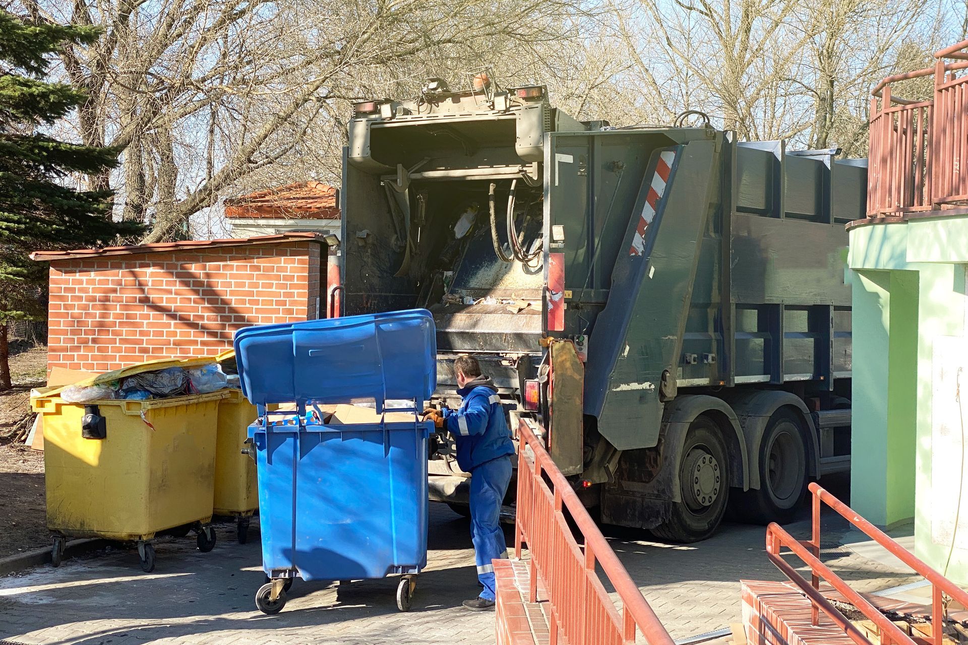 Garbage truck collecting blue and yellow bins beside a brick building on a street