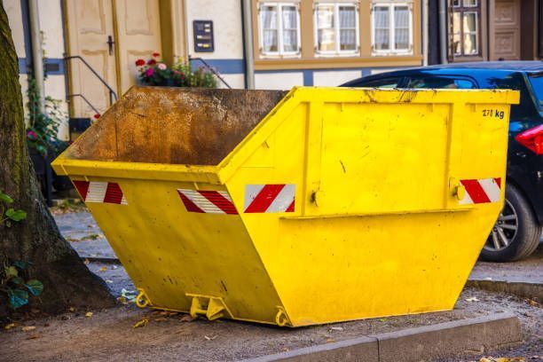 A yellow commercial skip bin with reflective stripes sits by a building and parked cars.