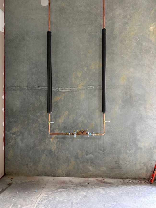 Copper plumbing pipes on a gray wall, forming a U-shape. Black insulation covers the vertical pipes.