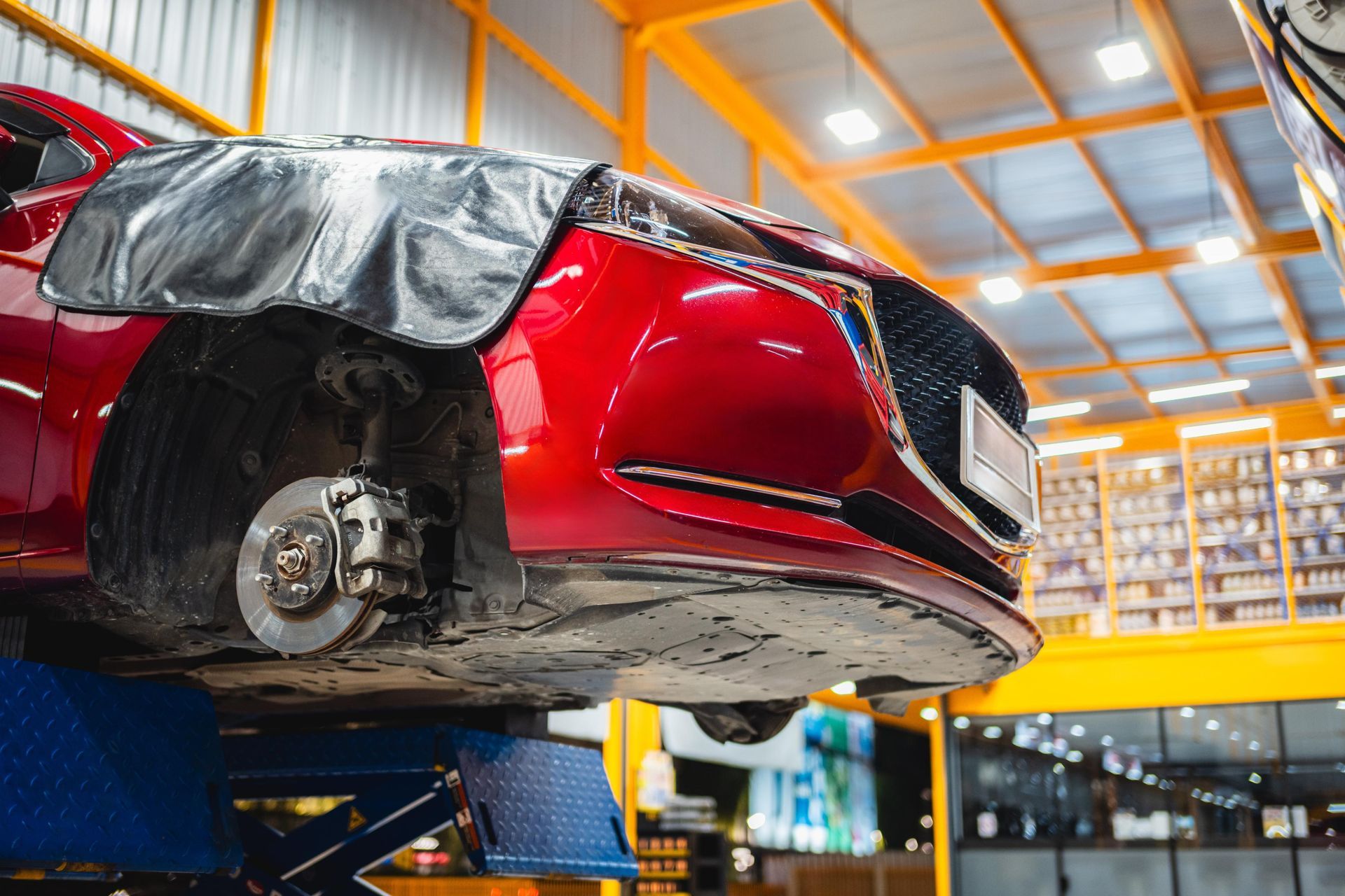 Red car body on a lift in an auto repair shop, with a wheel removed and rear panel covered in plastic.