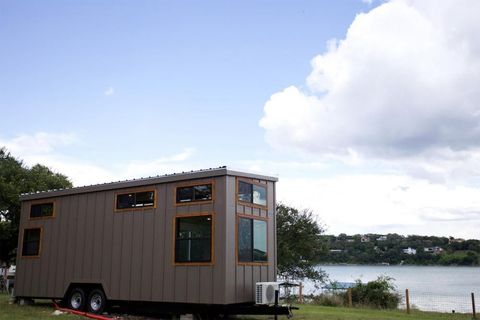 Tiny house on wheels near a lake, with brown siding, windows, and a cloudy sky.