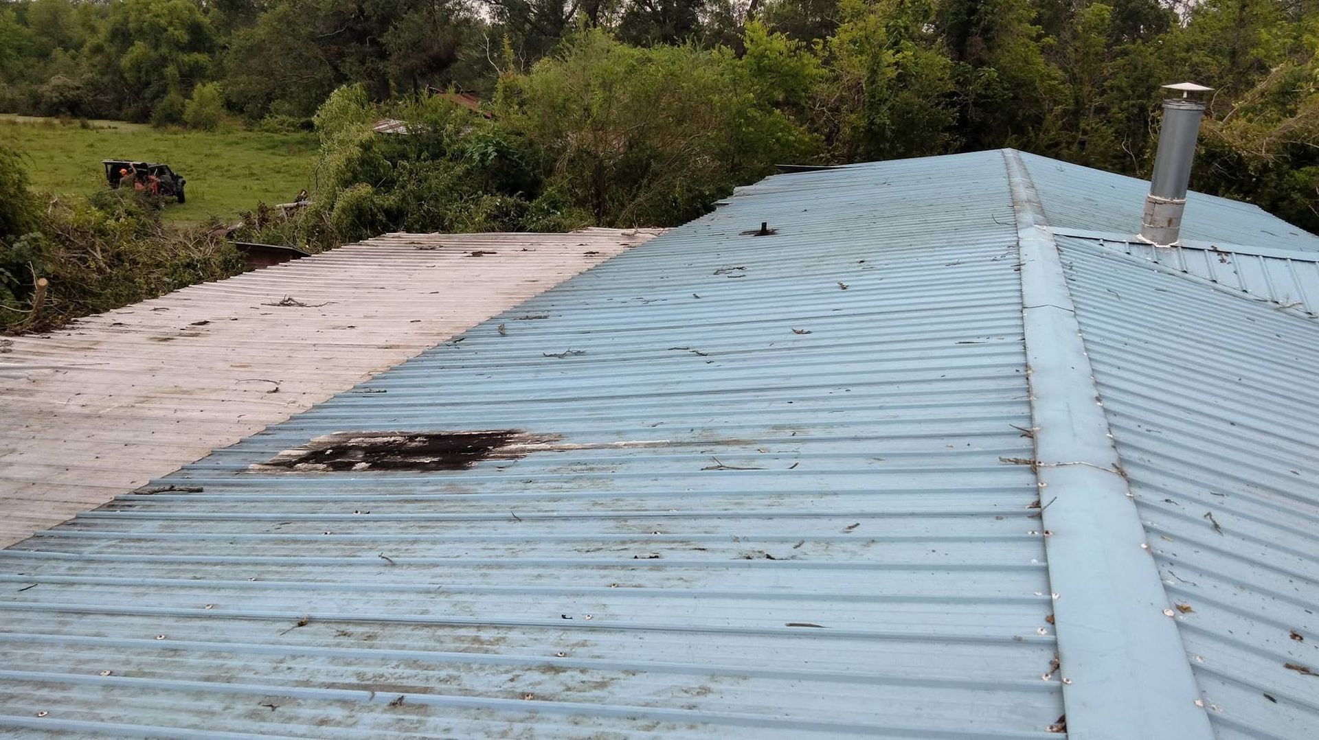 Blue corrugated metal roof with a damaged patch, surrounded by green trees and brush.