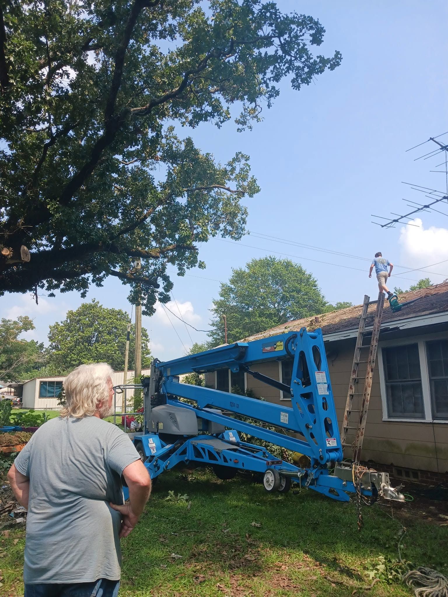 Blue boom lift collapsed against a house, with a person standing nearby in a yard.