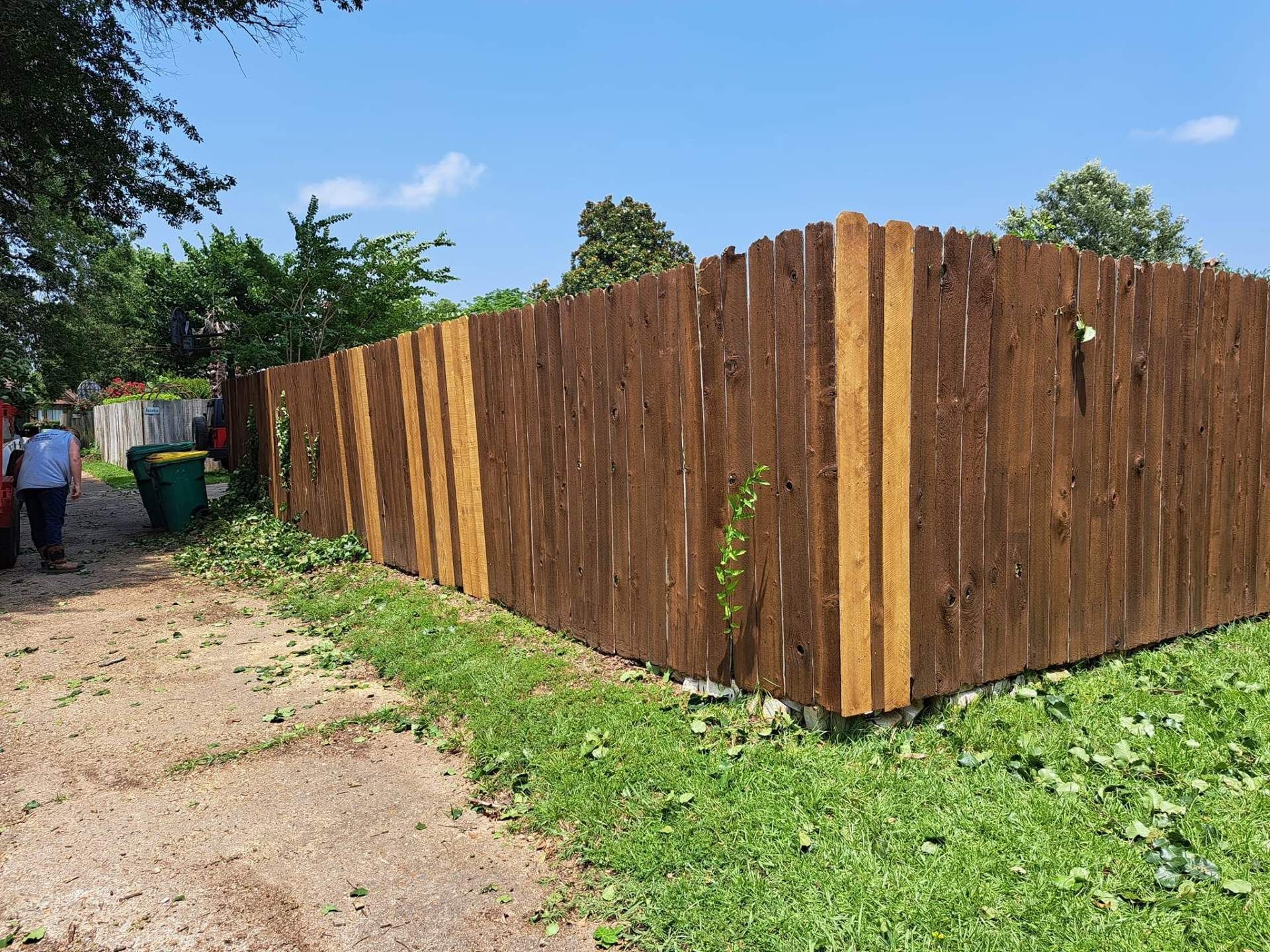 Wooden fence along a dirt path with greenery and people in the distance under a blue sky