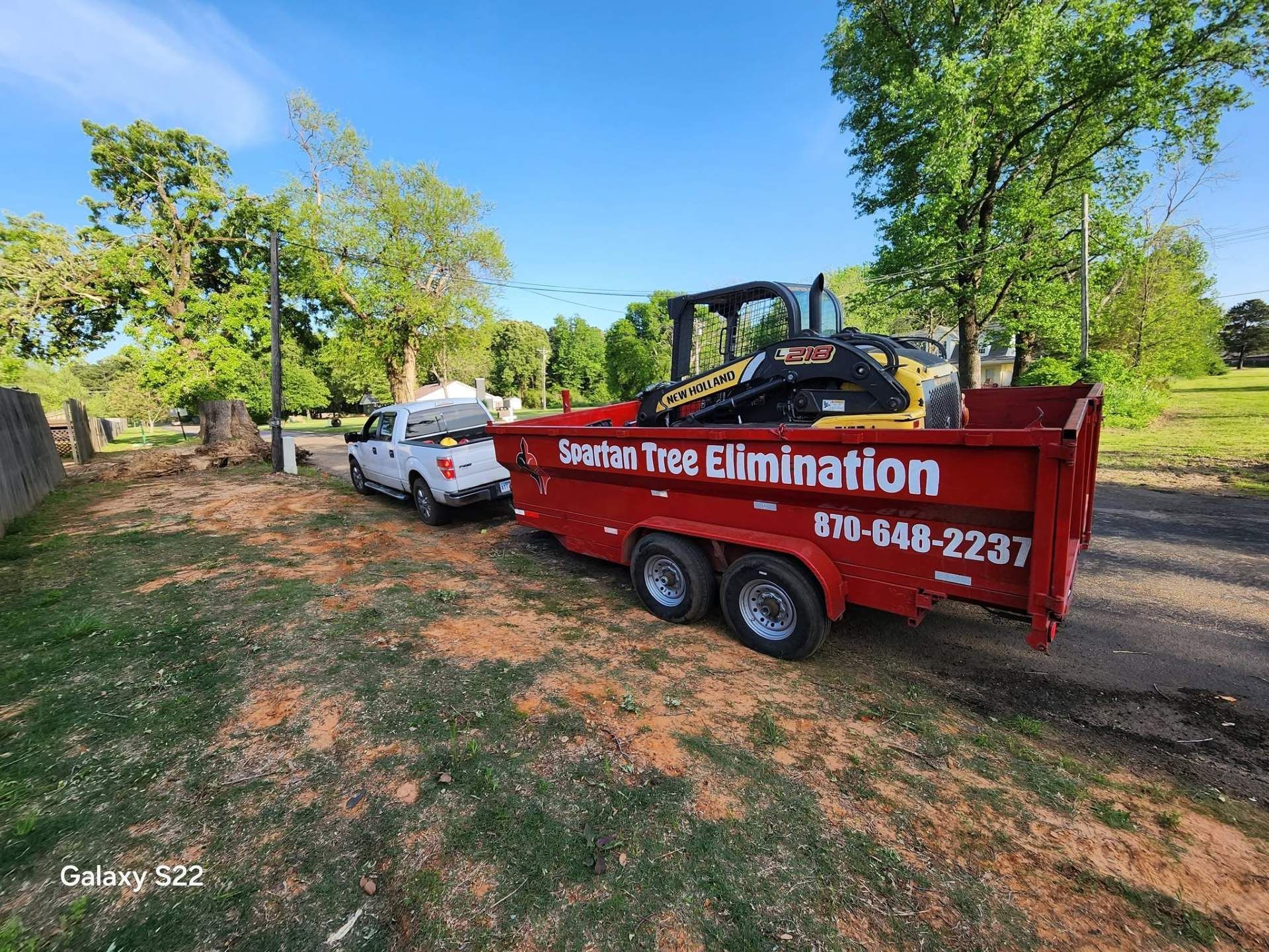 Red dump trailer labeled “Stump Tree Elimination” beside a white truck in a grassy yard with trees.