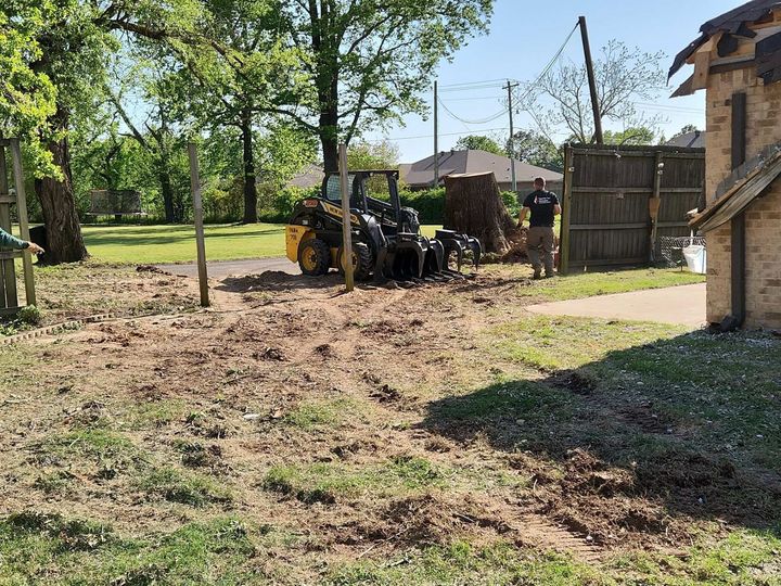 Yellow tractor in a muddy yard beside a house, with trees, a fence, and a person nearby