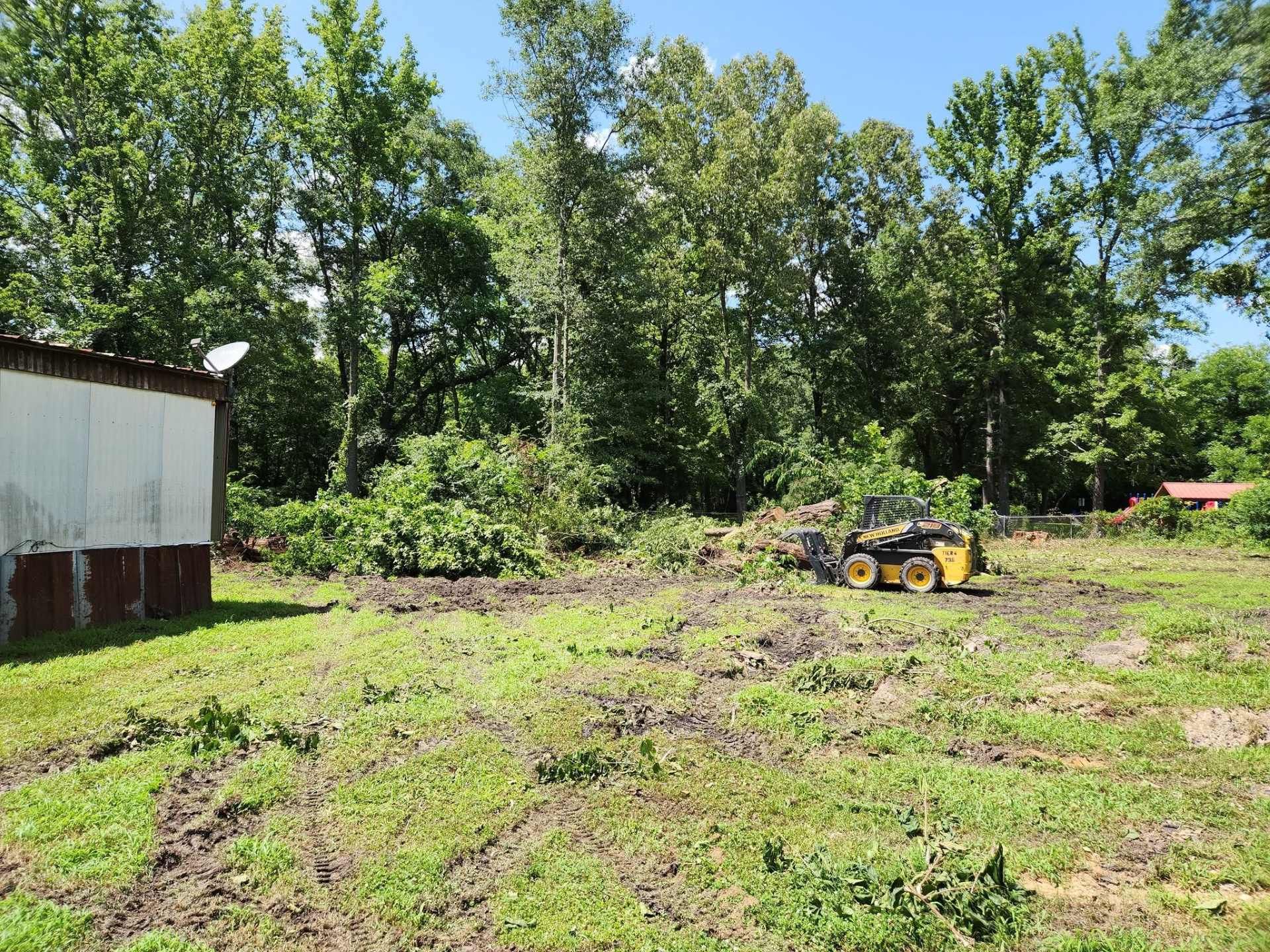 Small yellow tractor in a muddy field beside a white building and trees on a sunny day