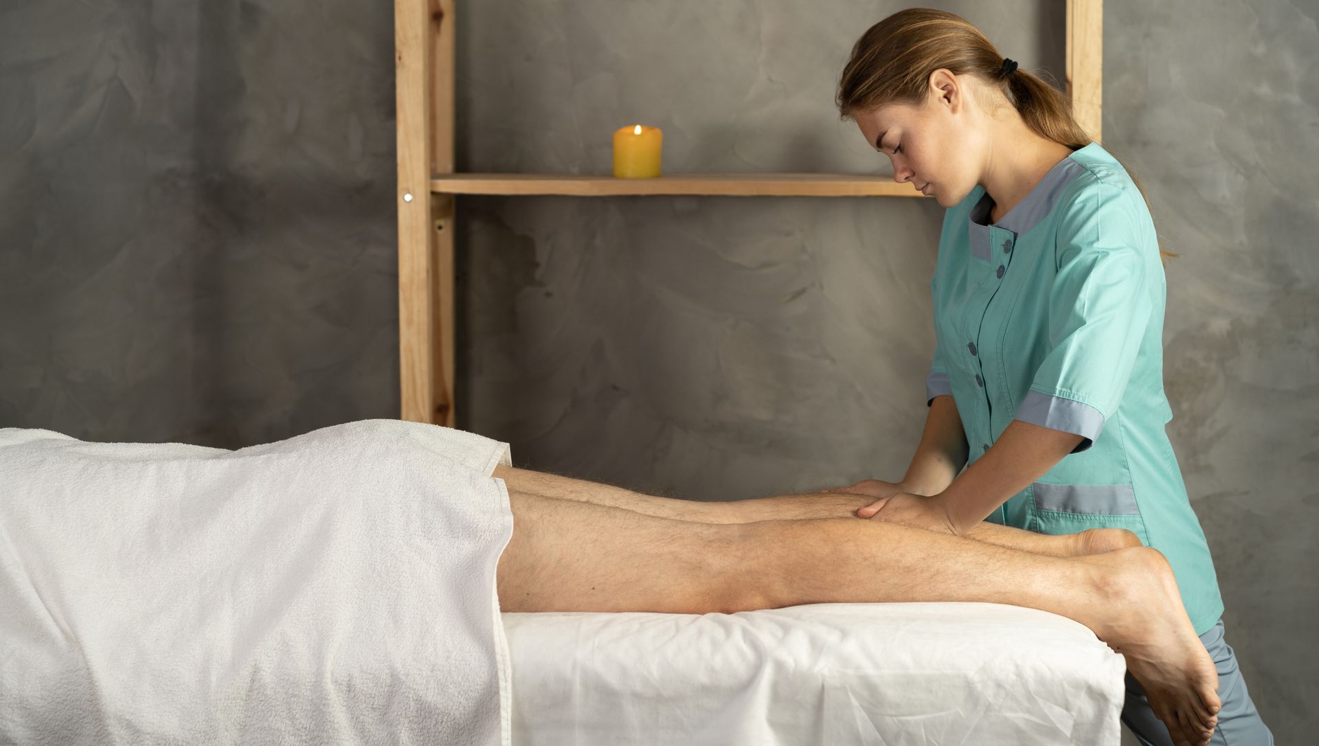 A massage therapist in a teal tunic massages the lower legs of a person lying on a spa table in a room with a candle.
