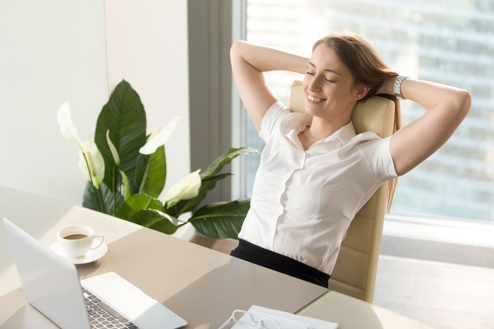 A woman is sitting at a desk with her hands behind her head.