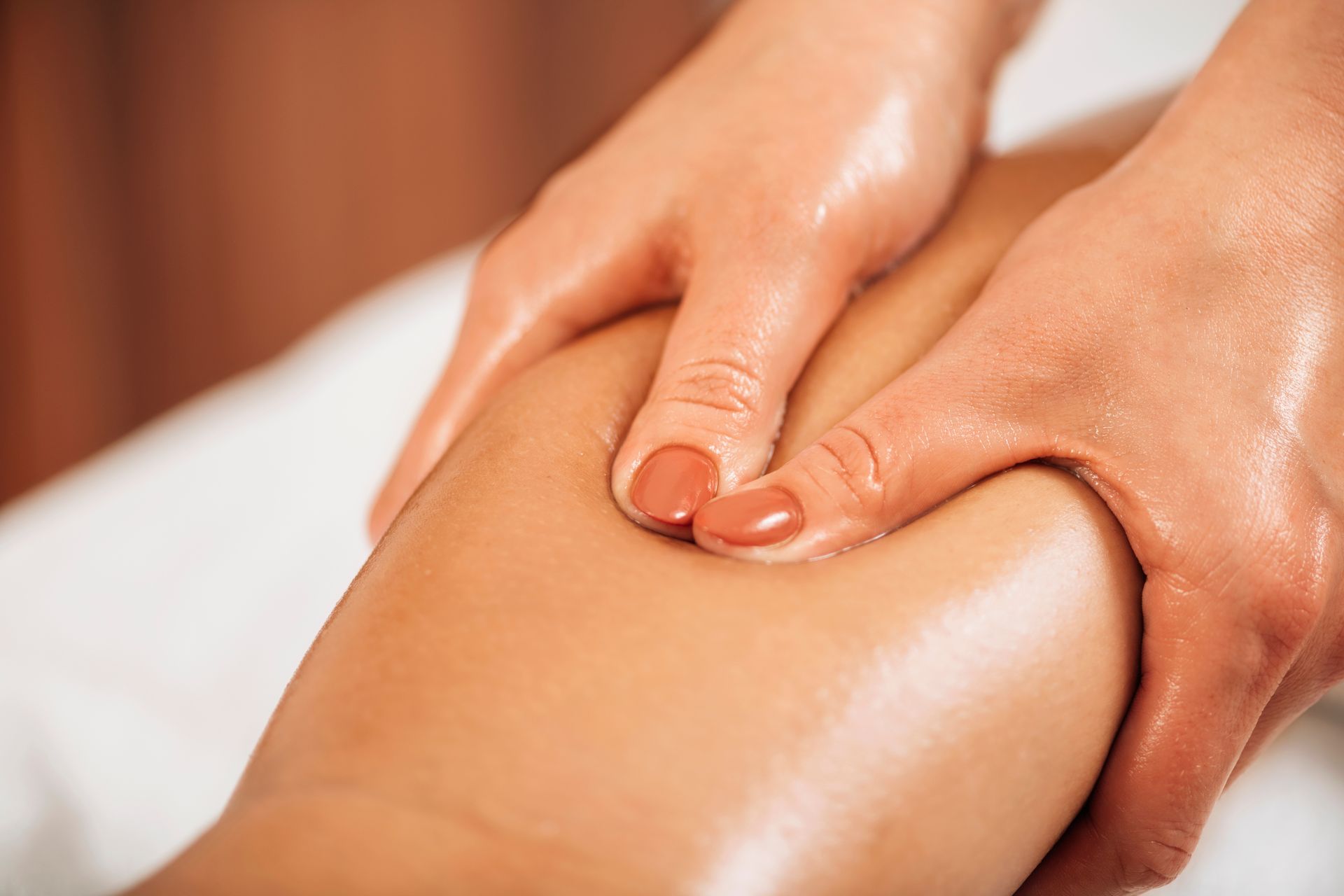 Hands massaging a leg with oil, on a white surface.