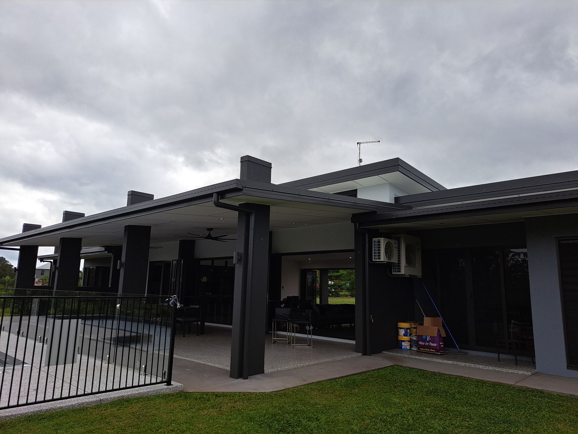 Black and gray modern house with a patio, columns, and a fence, under a cloudy sky.
