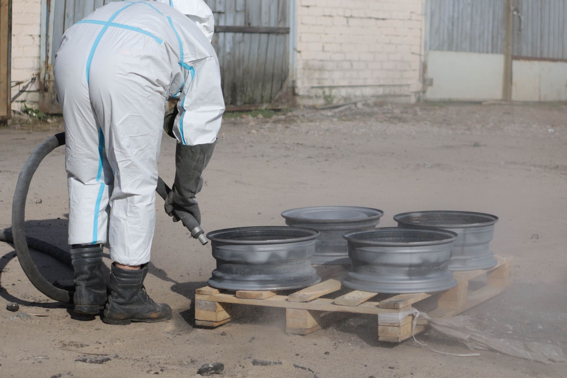 Worker in white protective suit blasting dust from stacked metal wheels with an air hose outdoors