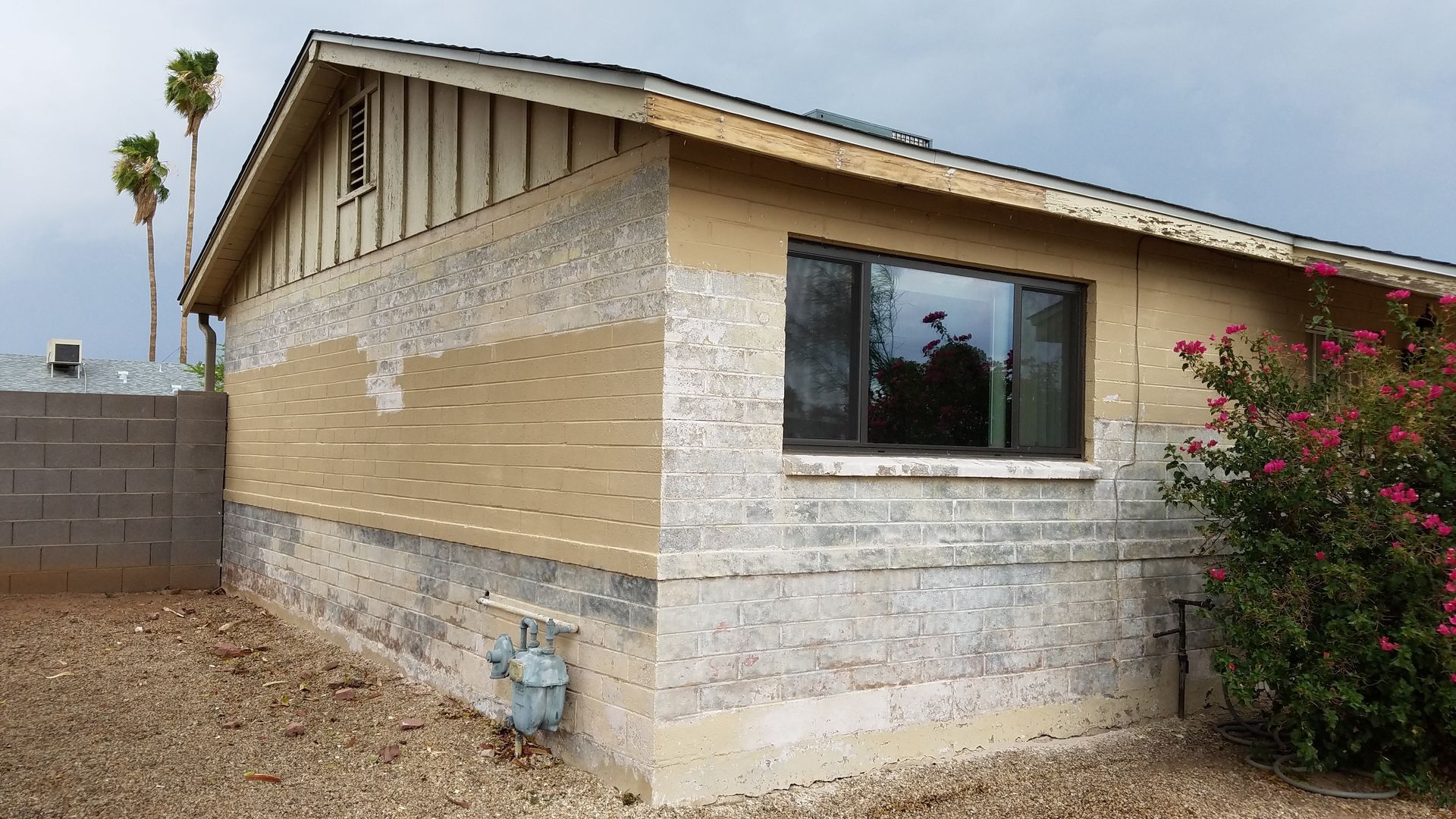 Single-story beige house exterior with a large window, gravel yard, and flowering shrub. Single-story beige house exterior with a large window, gravel yard, and flowering shrub.