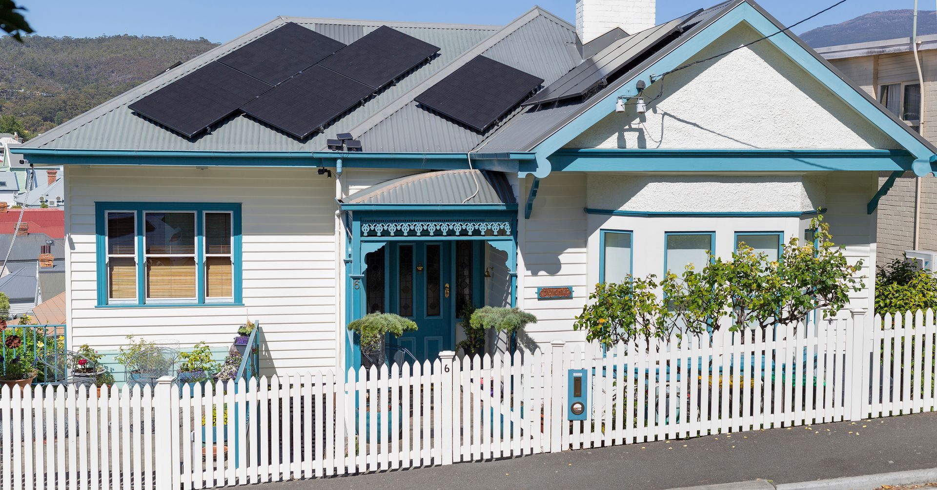 White house with teal trim, solar panels, and a white picket fence in a sunny suburban neighborhood