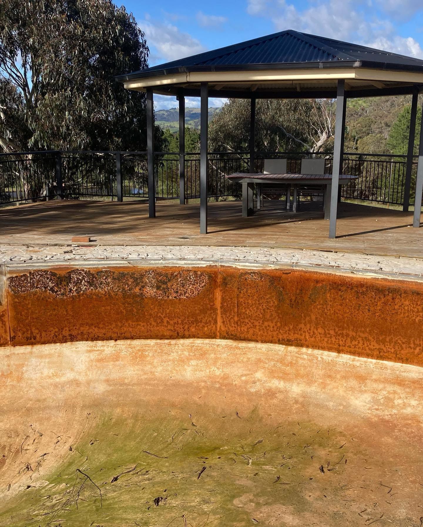 Empty concrete pool beside a gazebo with tables in a fenced outdoor area.