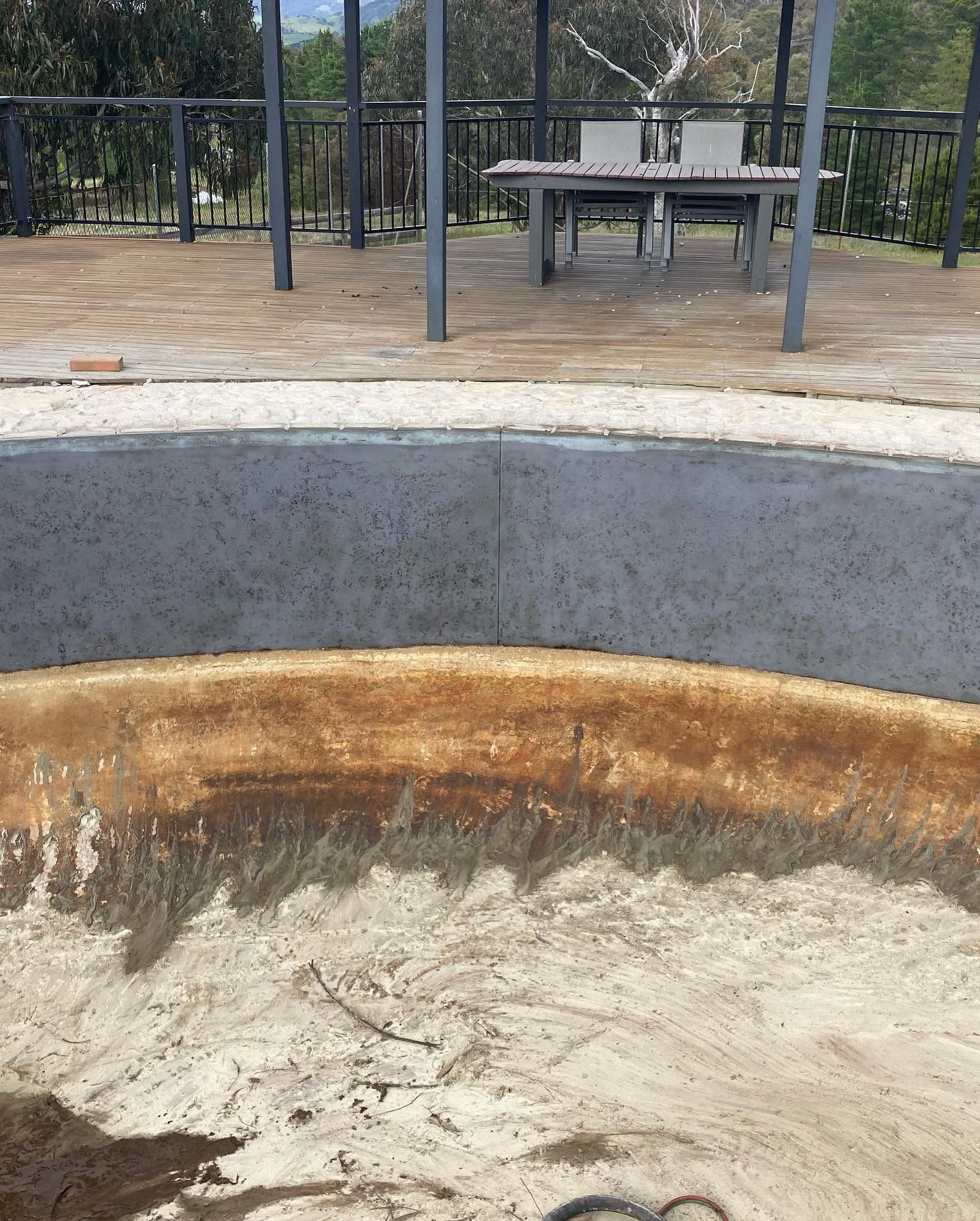 Empty concrete fire pit with rust stains in the foreground, with a picnic table and railings in the background