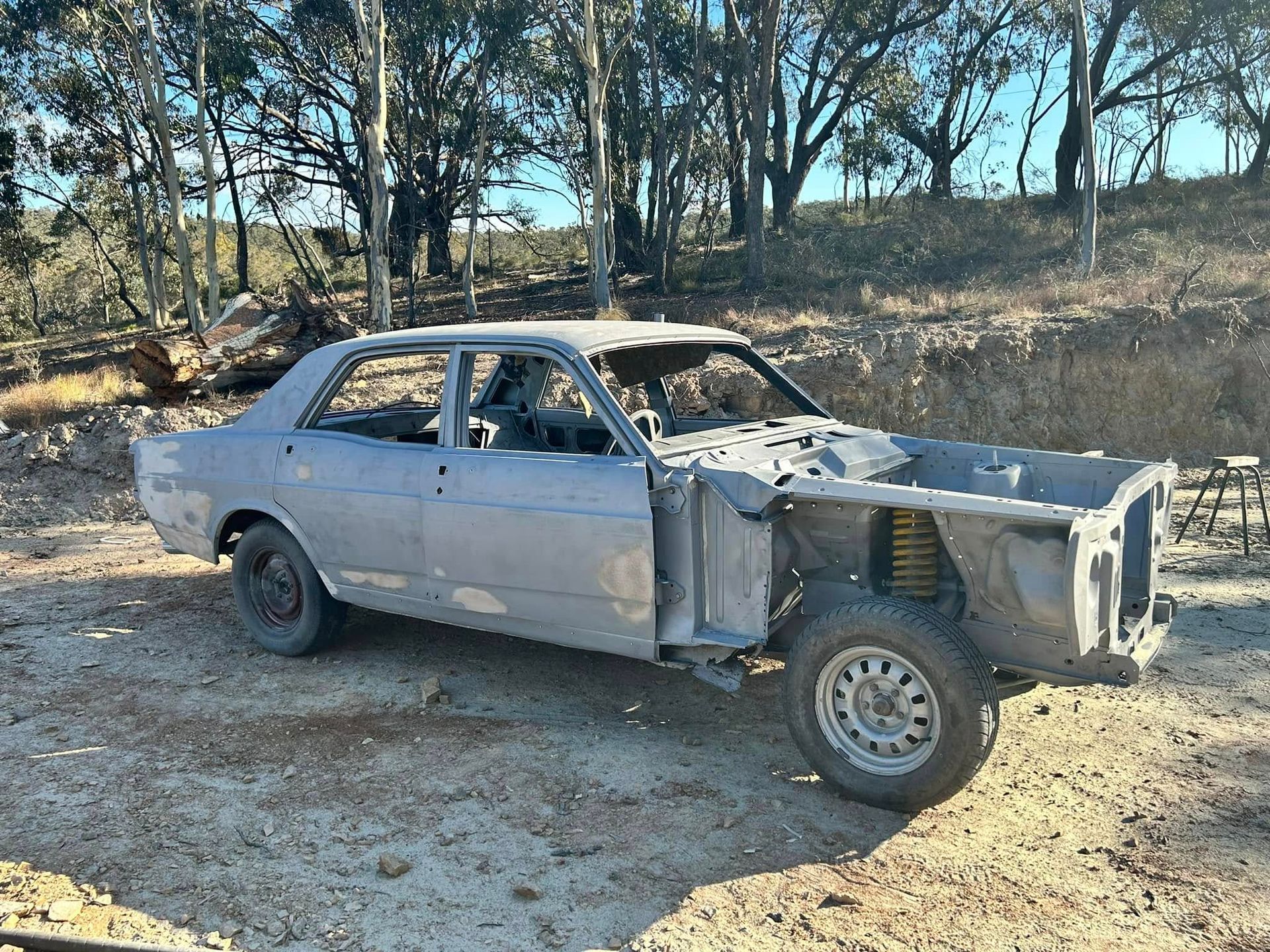 Damaged silver sedan with missing front end parked on a dirt road near trees and dry brush