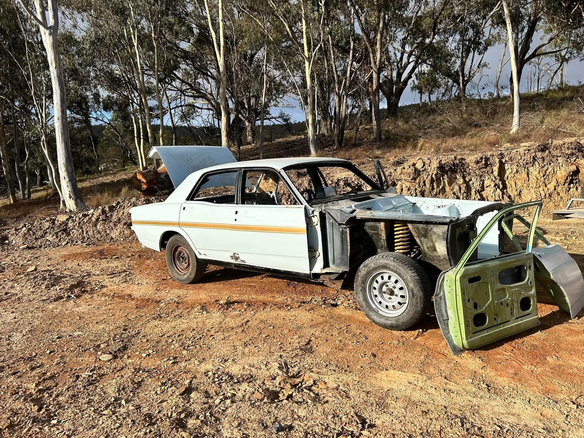 Damaged white car with detached green front end on a dirt road in a wooded area
