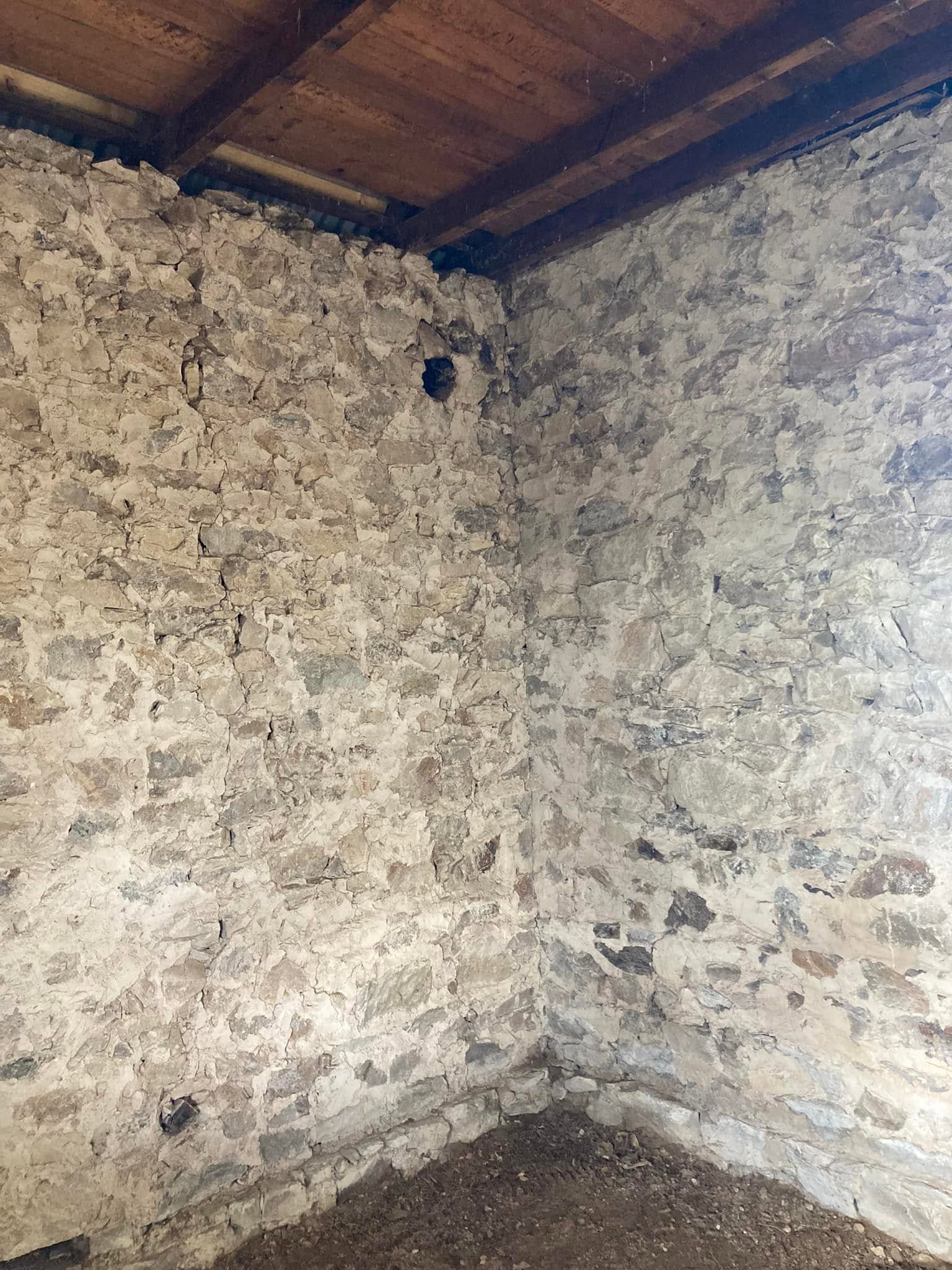 Empty stone room corner with rough white walls and a wooden ceiling, with a small hole near the top.