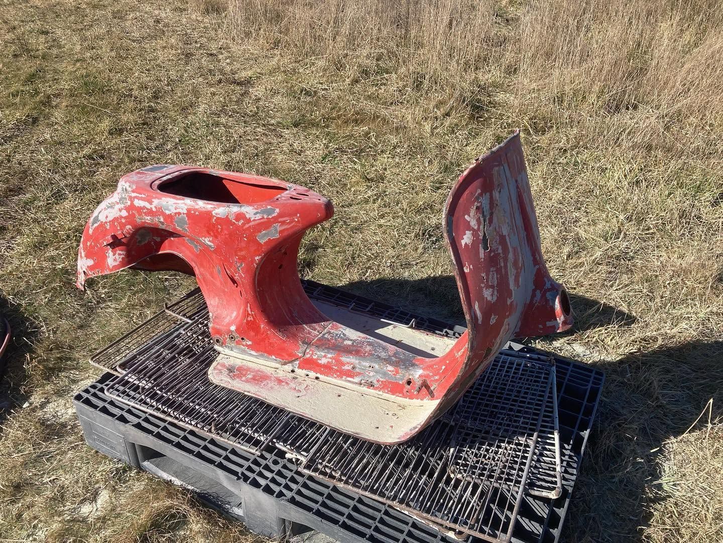 Red damaged scooter body panel sitting on a black metal grate outdoors on dry grass.