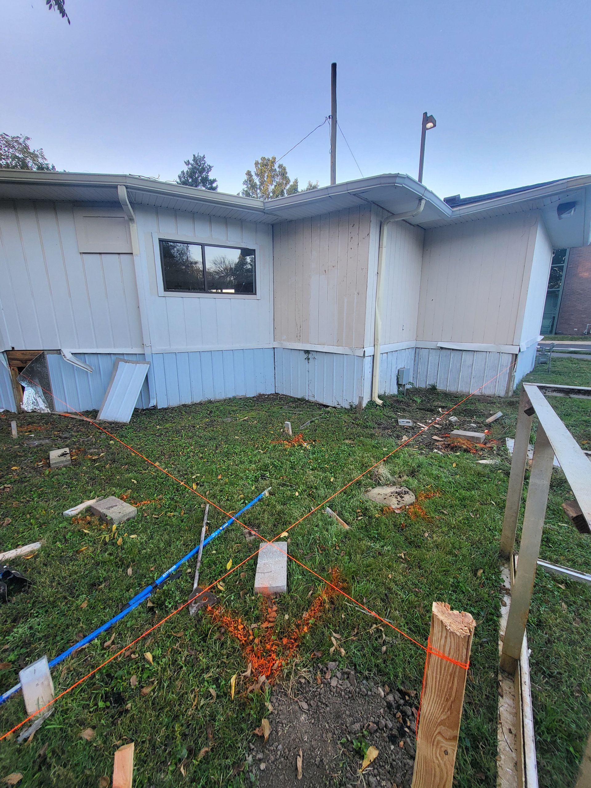 A light-colored house exterior with sections of siding missing, set near a lawn marked by string lines and wooden stakes.