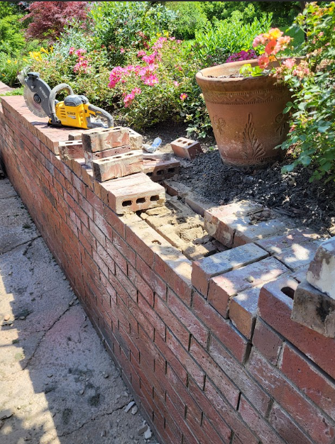 A power saw sits atop a brick garden wall undergoing repairs, with loose bricks, mortar, and a flower pot nearby.