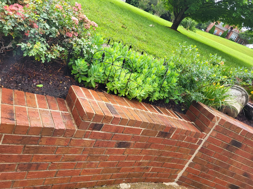 A multi-tiered red brick retaining wall borders a garden bed with green shrubs and rose bushes against a grassy lawn.