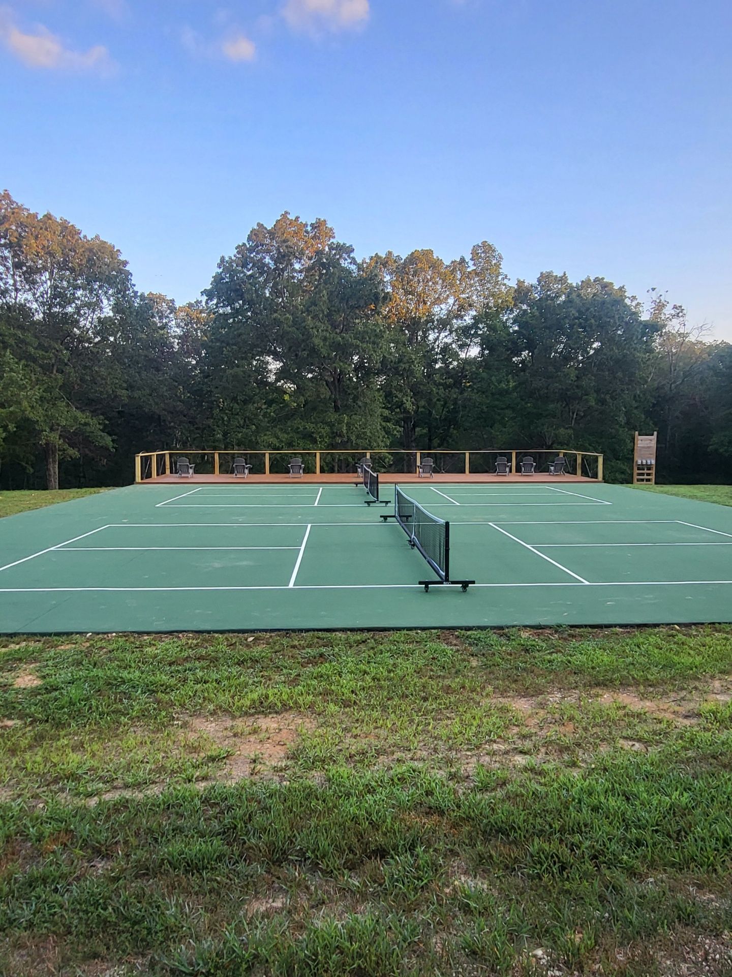 A green pickleball court outdoors, featuring a black net, white court lines, and a wooden viewing deck in the background.