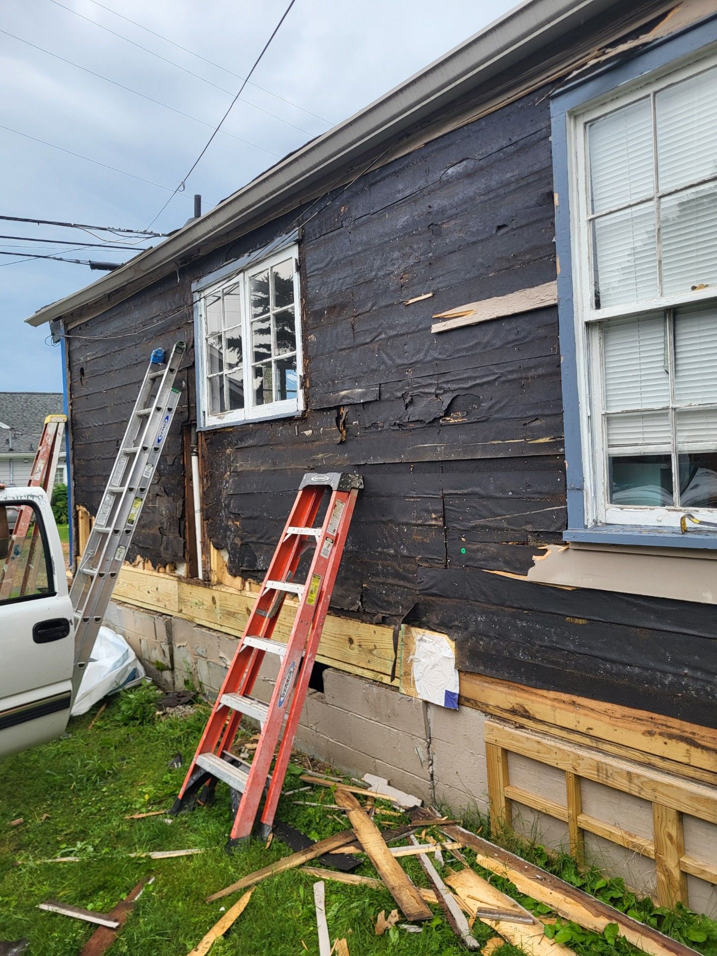 Exterior view of a house during siding removal, showing exposed dark sheathing and two ladders against the wall.