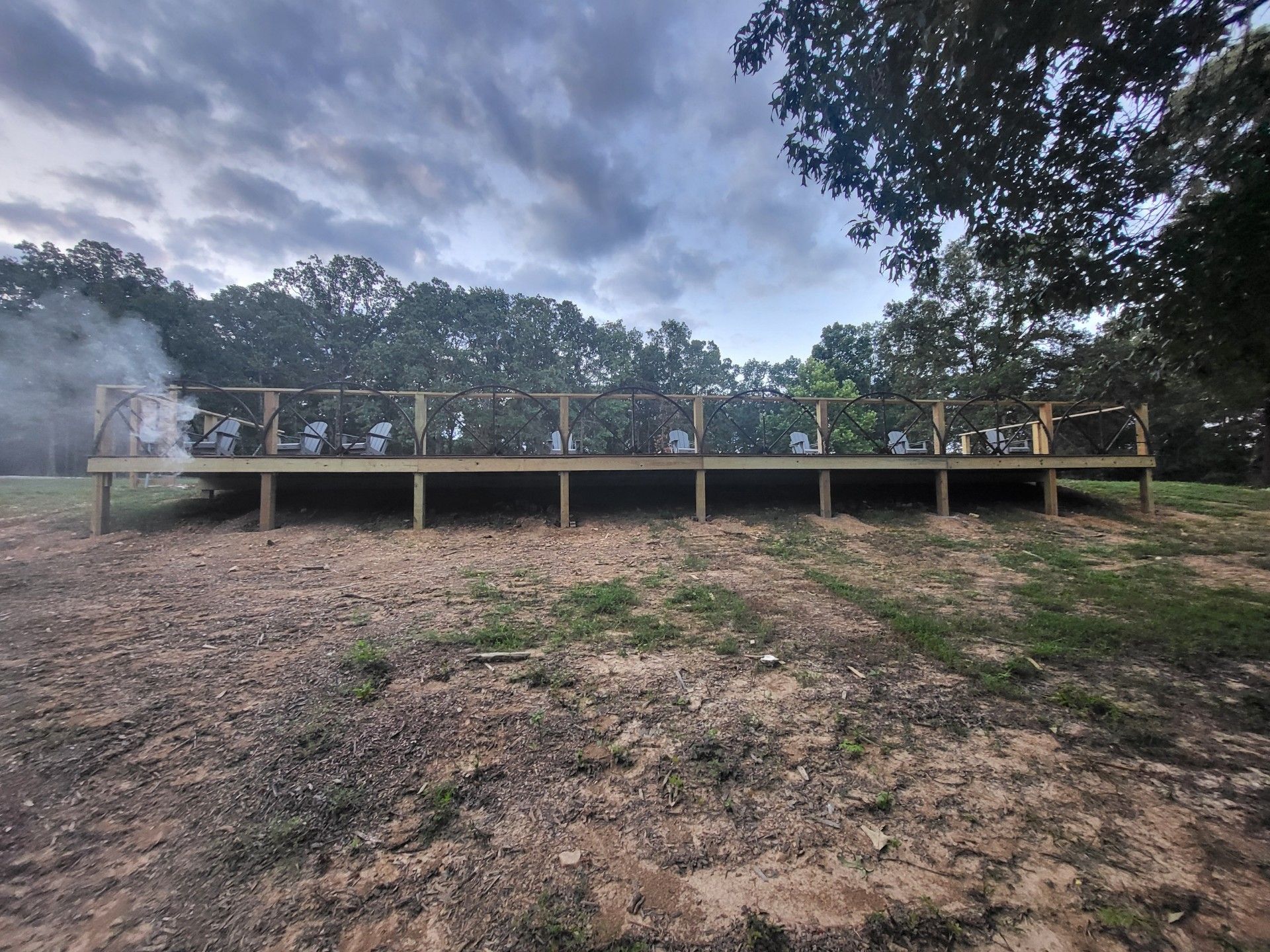 A low wooden deck with railings stands on bare ground before a line of trees under a cloudy, overcast sky.
