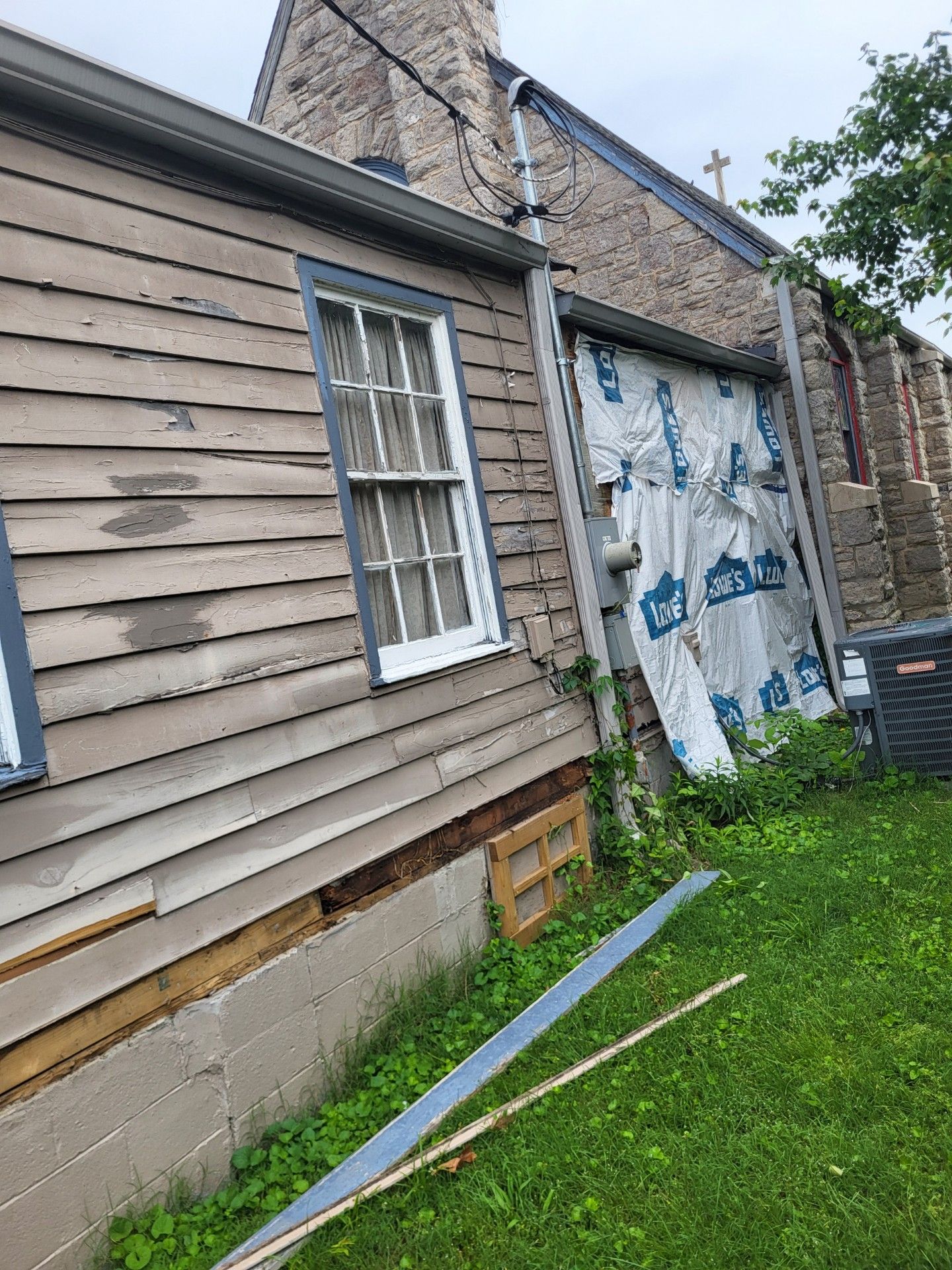 A view of a weathered building with wood siding, a stone wall section covered by a protective tarp, and a grass yard.