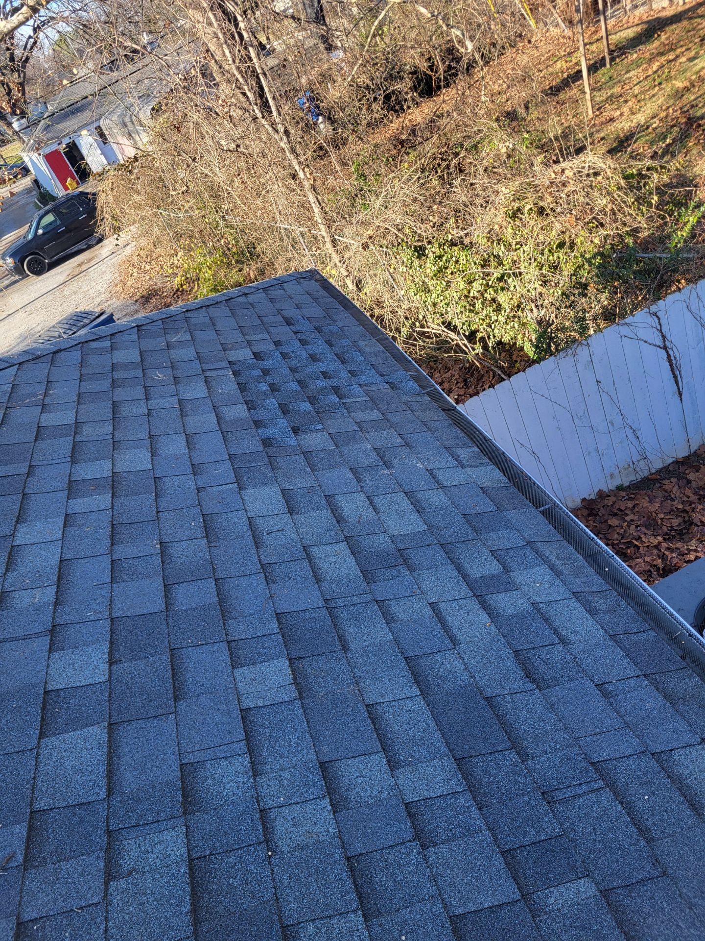 A high-angle view looking down at a gray asphalt shingled roof sloping toward a tree-filled backyard.