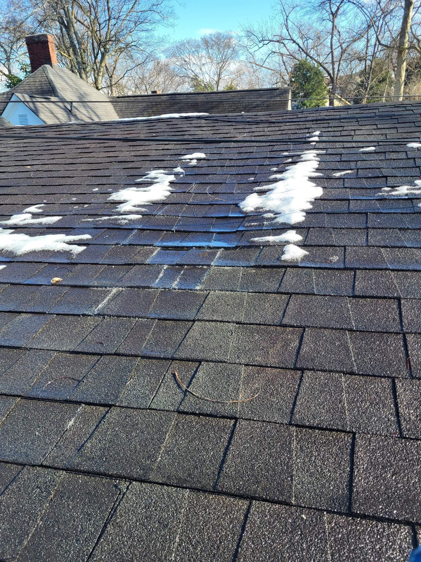Patches of snow melting on a dark, textured asphalt shingle roof on a sunny day.