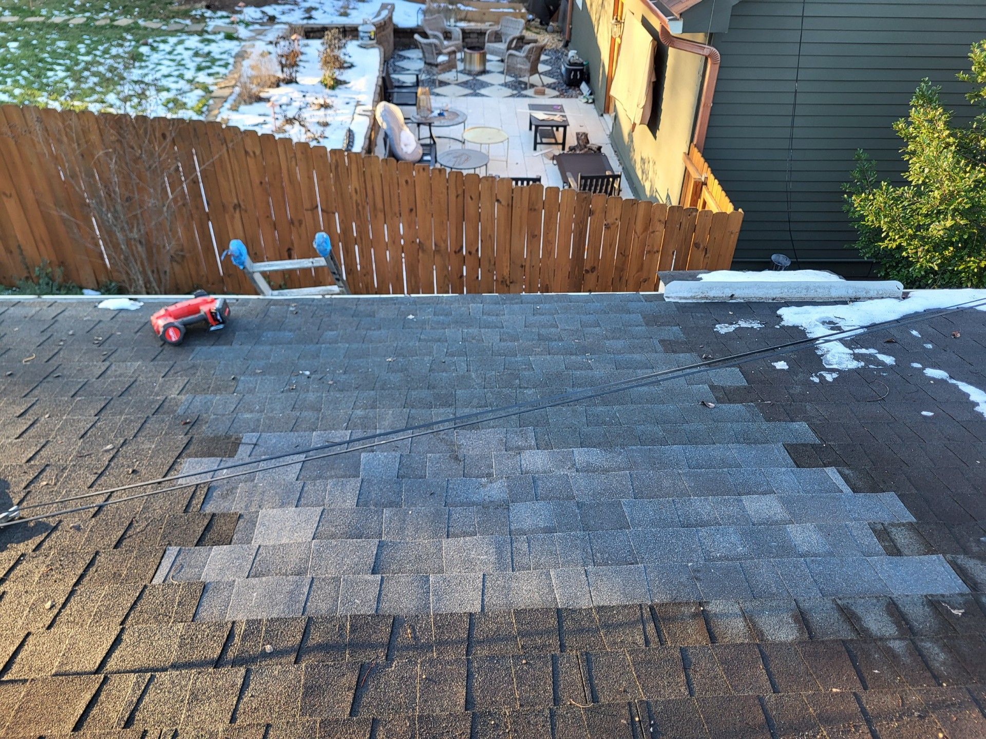 A patch of new, lighter grey shingles on a weathered dark roof, viewed from above near a backyard fence.