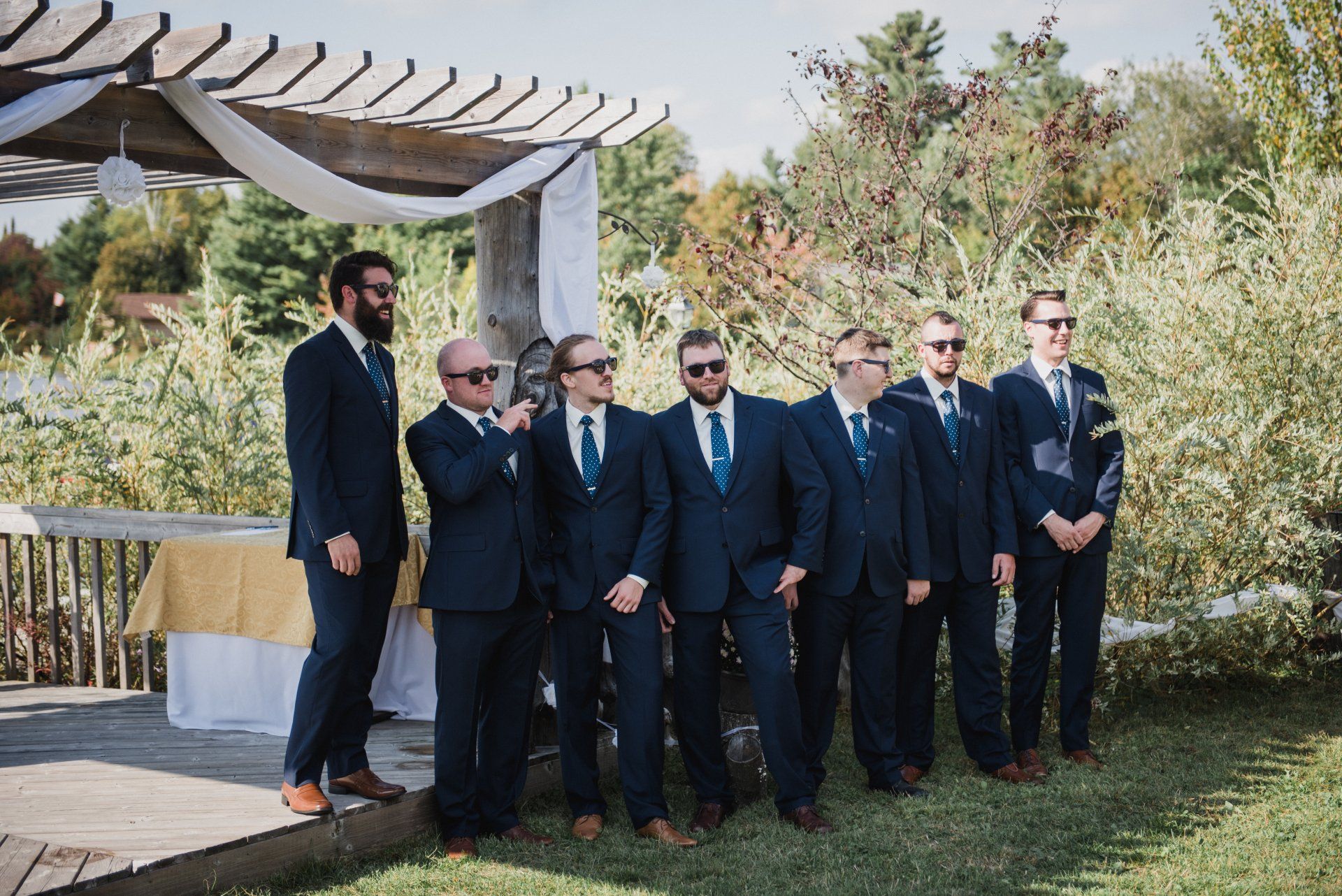 Seven men in dark suits stand together outdoors under a white pergola, posing for a group photo.
