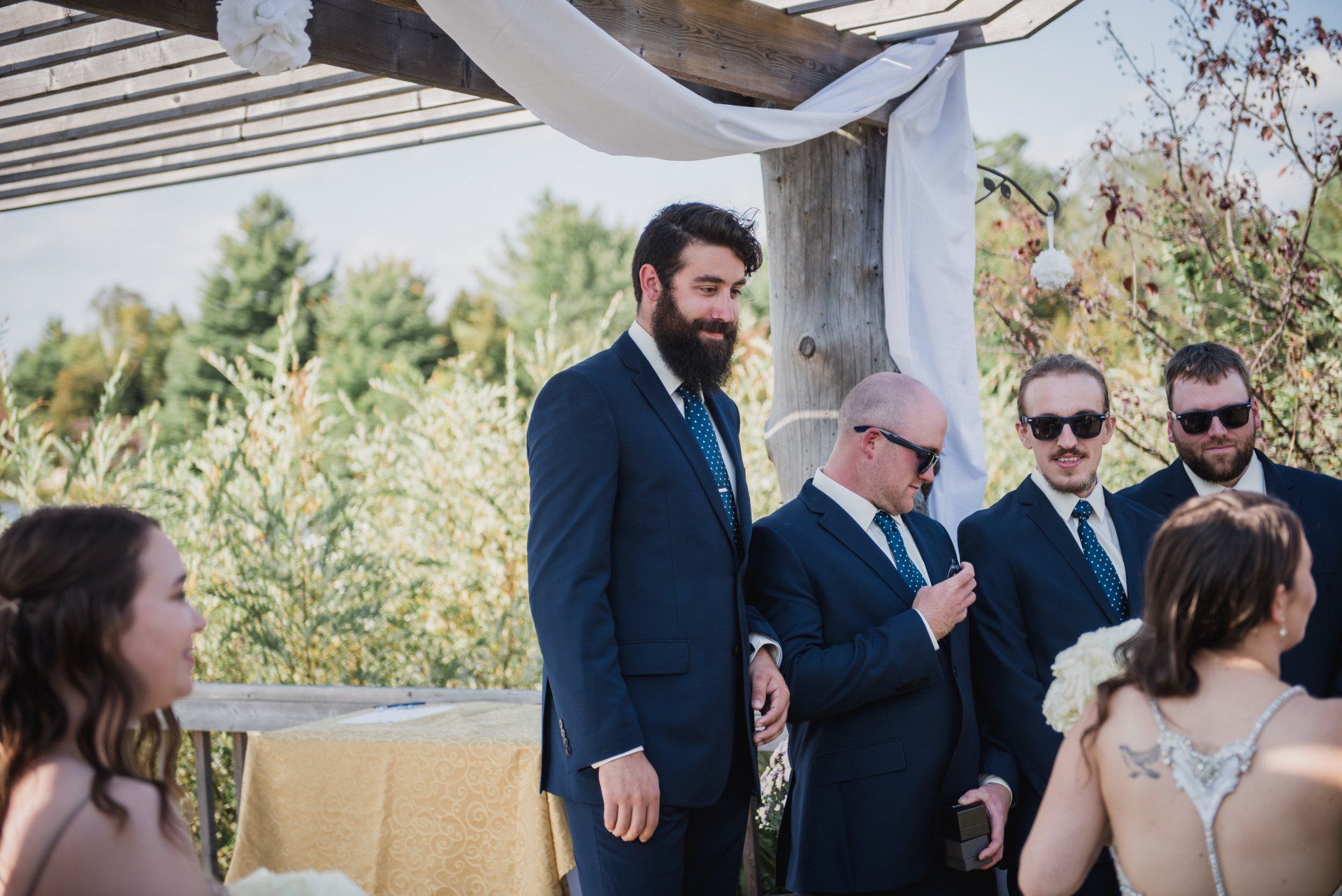Guests stand under a canopy at an outdoor wedding ceremony, with a bearded man in a navy suit centered.