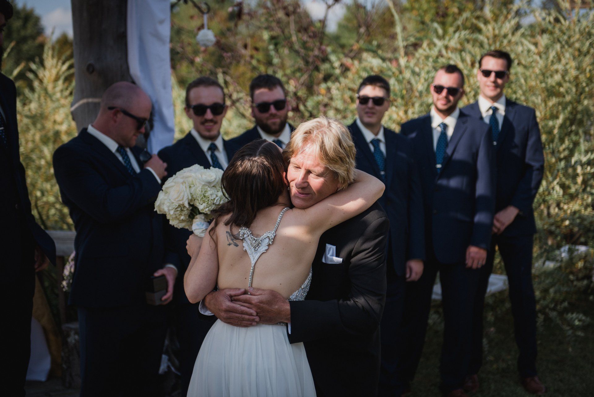 Wedding guests embrace outdoors beside the bridal party in suits and sunglasses.