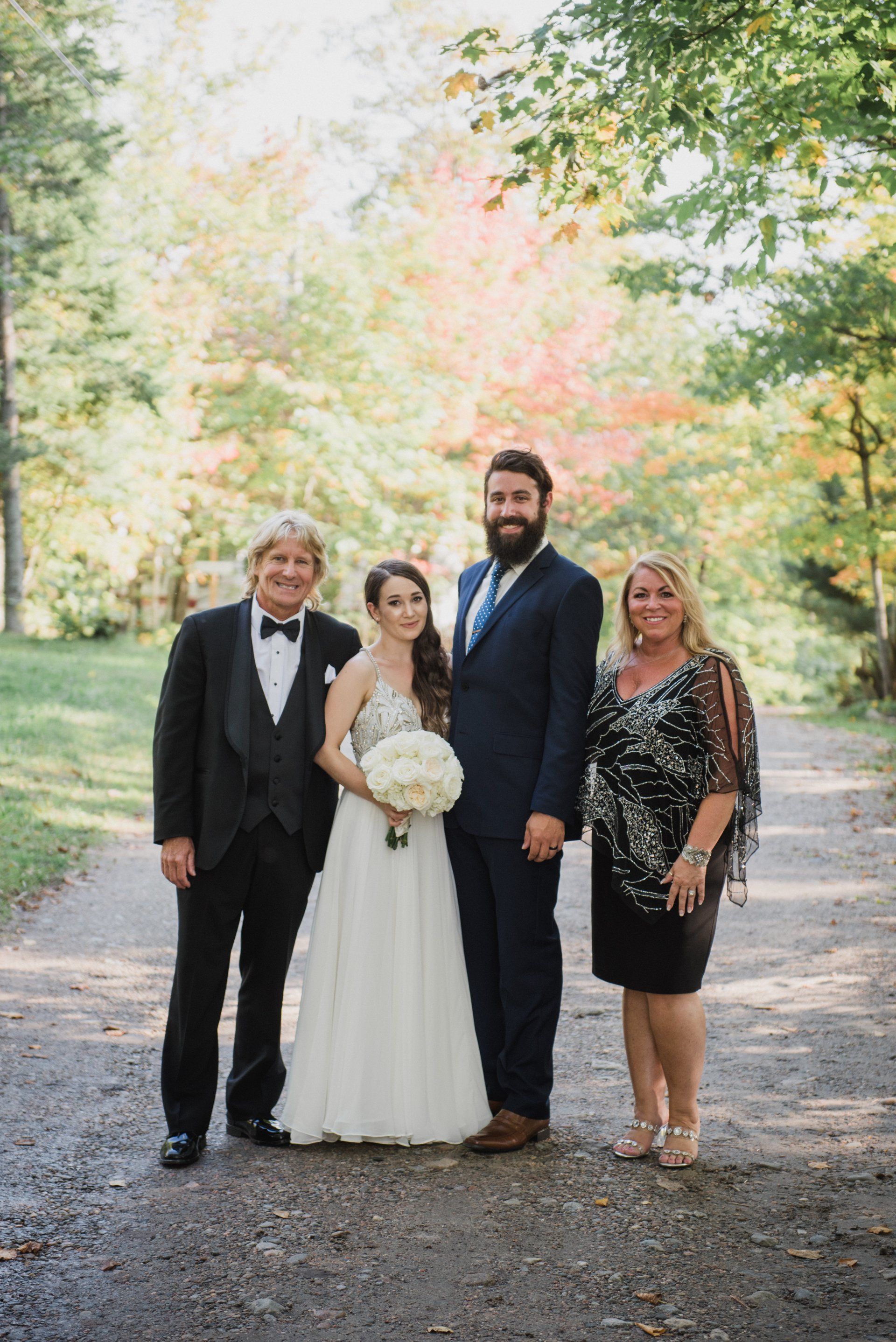 Four people standing on a tree-lined path at an outdoor wedding, with the bride holding a bouquet.